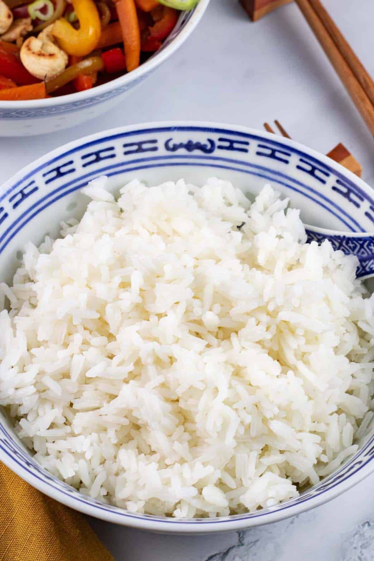 Steamed jasmine rice in a blue and white oriental bowl with spoon, chopsticks and a bowl of chicken and cashew nut stir-fry on the side.
