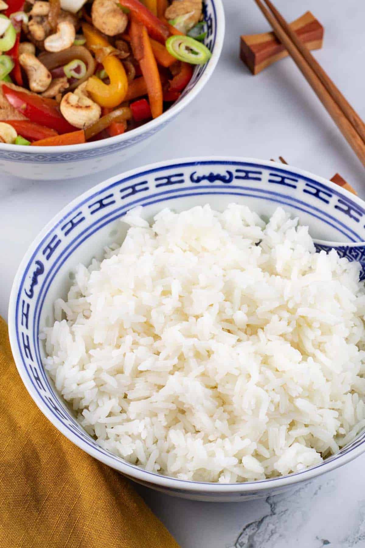 Steamed jasmine rice in a blue and white oriental bowl with spoon, chopsticks and a bowl of chicken and cashew nut stir-fry on the side.