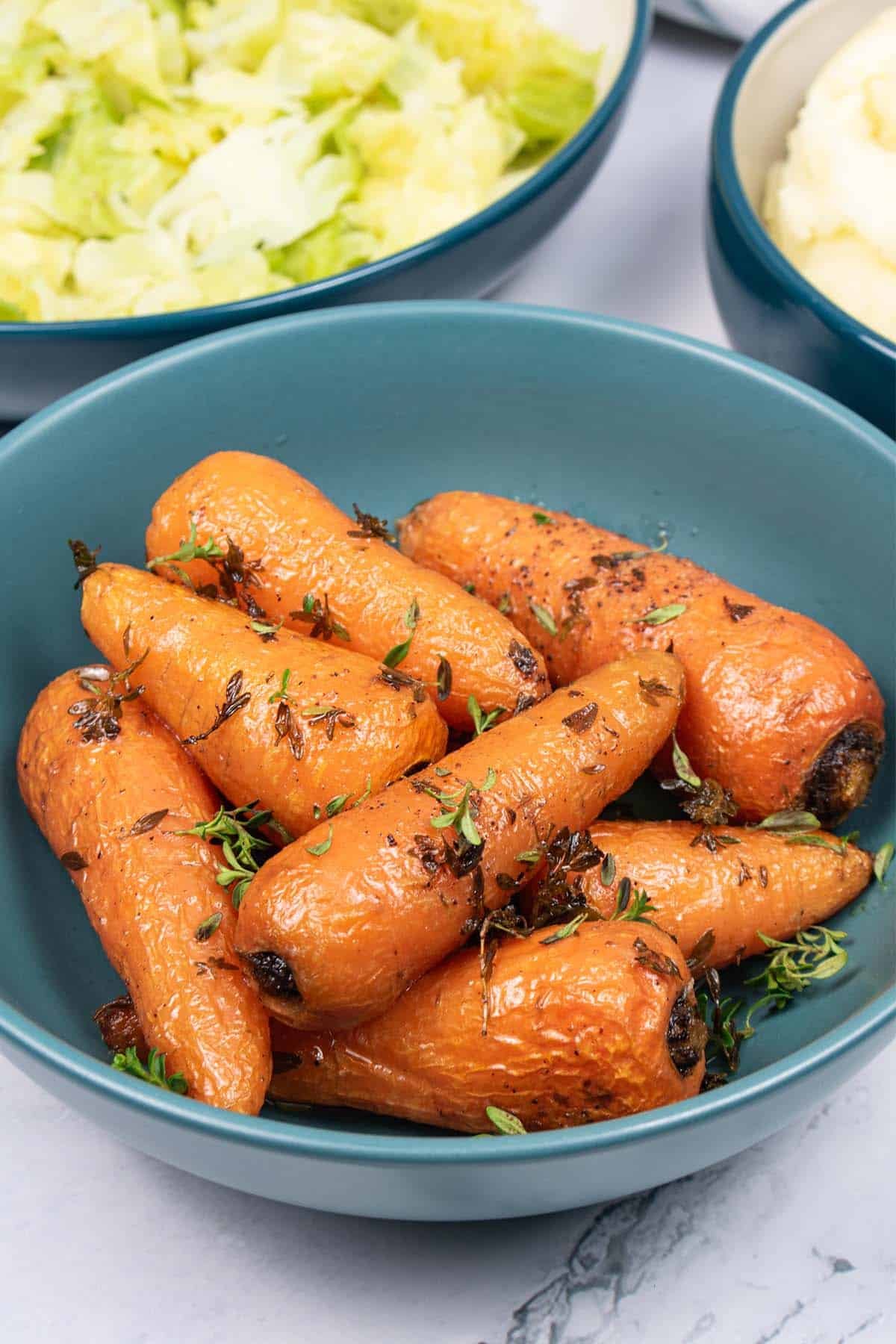 Thyme butter roasted Chantenay carrots in a teal bowl, with bowls of cabbage and mashed potatoes in the background.