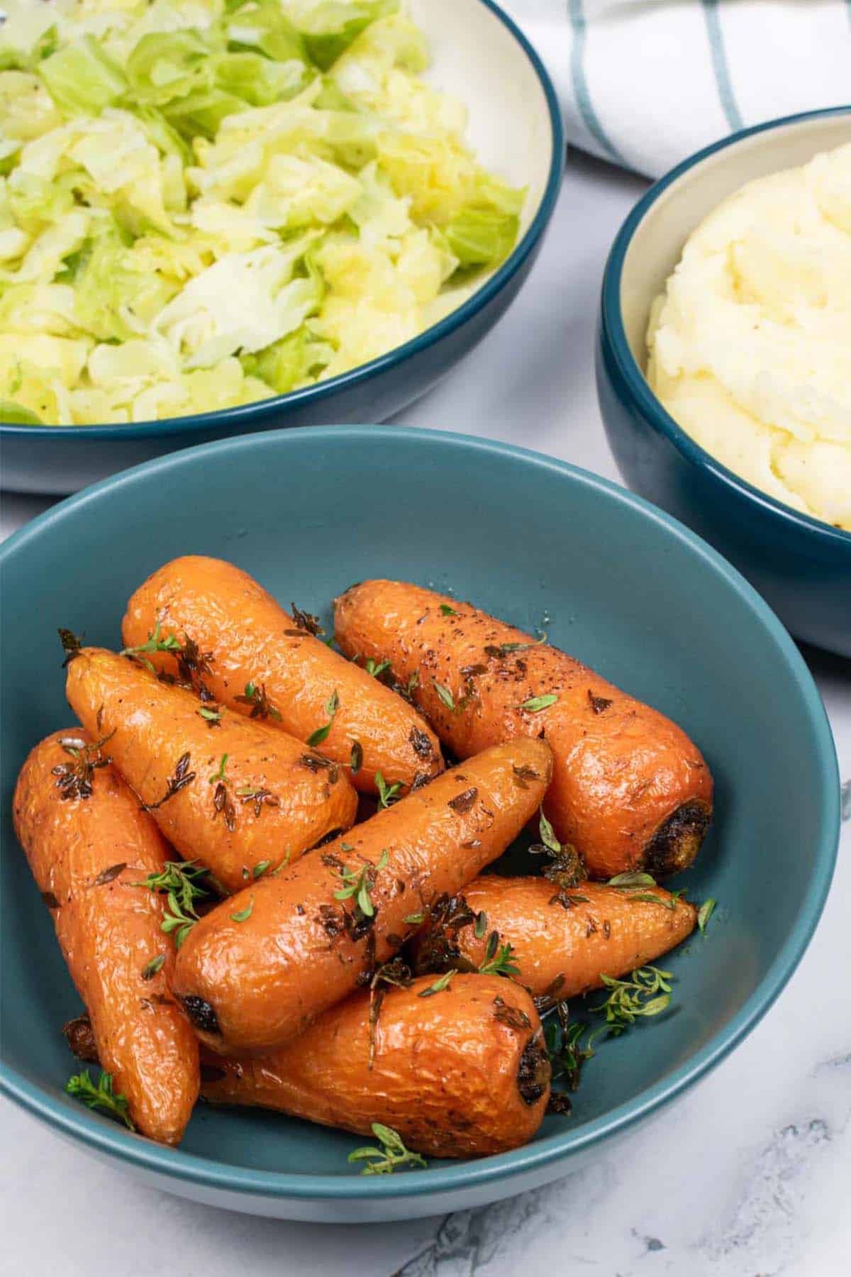 Thyme butter roasted Chantenay carrots in a teal bowl, with bowls of cabbage and mashed potatoes in the background.