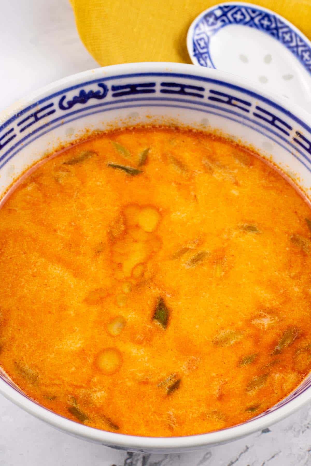 Tom yum soup in a small black blue and white oriental bowl with a spoon and mustard napkin on the side.