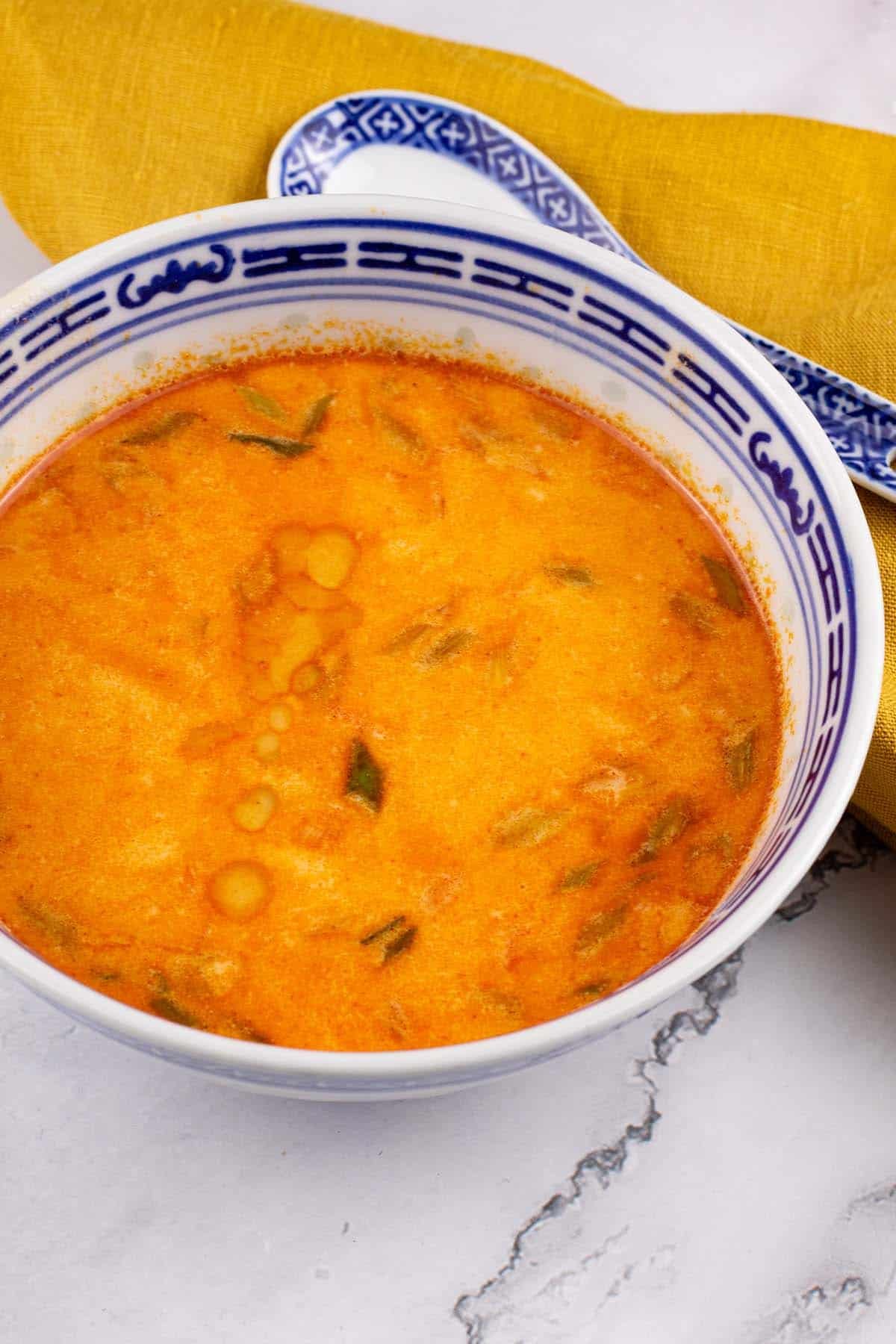 Tom yum soup in a small black blue and white oriental bowl with a spoon and mustard napkin on the side.