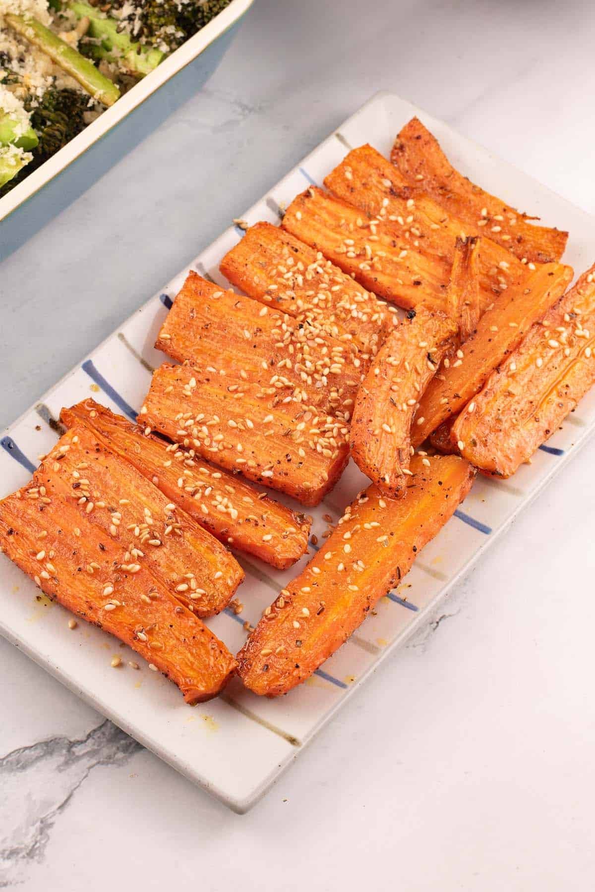 Za'atar roasted carrots on a rectangle striped platter with a dish of tenderstem broccoli on the side.