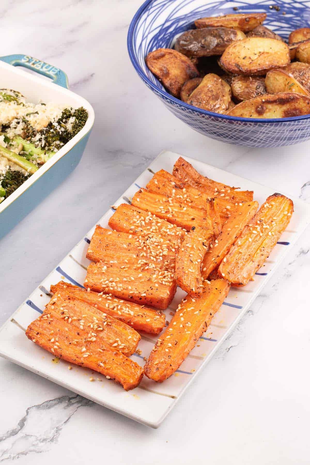 Za'atar roasted carrots on a rectangle striped platter with a dish of tenderstem broccoli and bowl of sautéed potatoes on the side.