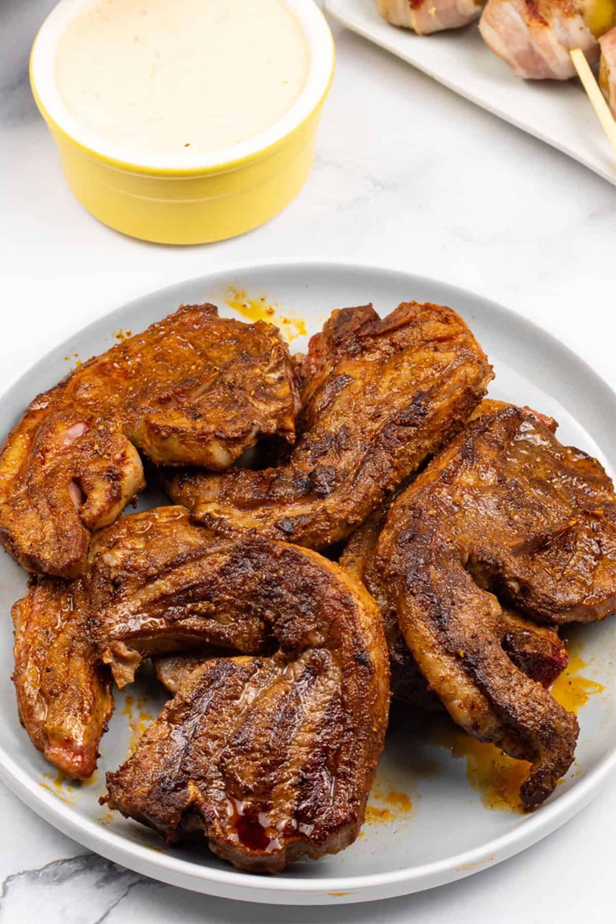 BBQ merguez lamb chops on a grey plate and a plate of baby new potatoes wrapped in bacon with harissa dip on the side.