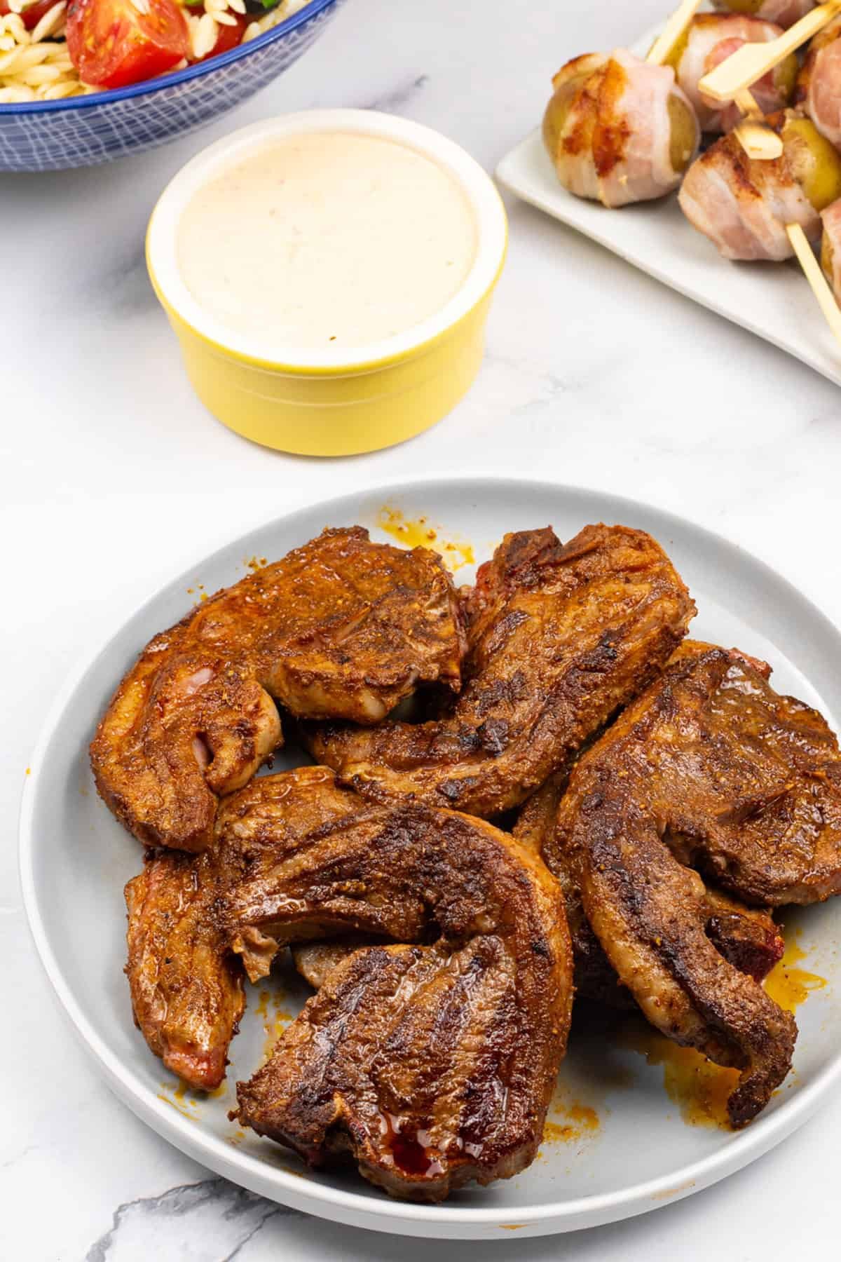 BBQ merguez lamb chops on a grey plate with a bowl of orzo salad with feta, and a plate of baby new potatoes wrapped in bacon with harissa dip on the side.