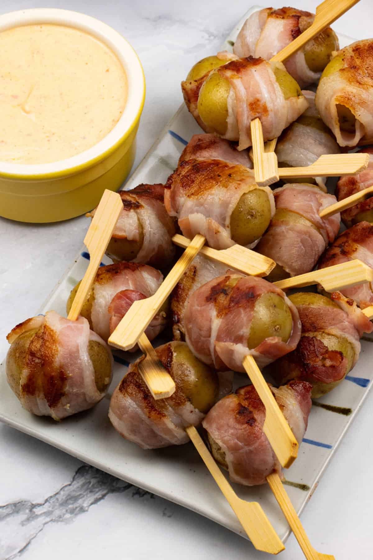 Baby new potatoes wrapped in bacon on a striped platter with harissa cheese dip in a yellow ramekin.
