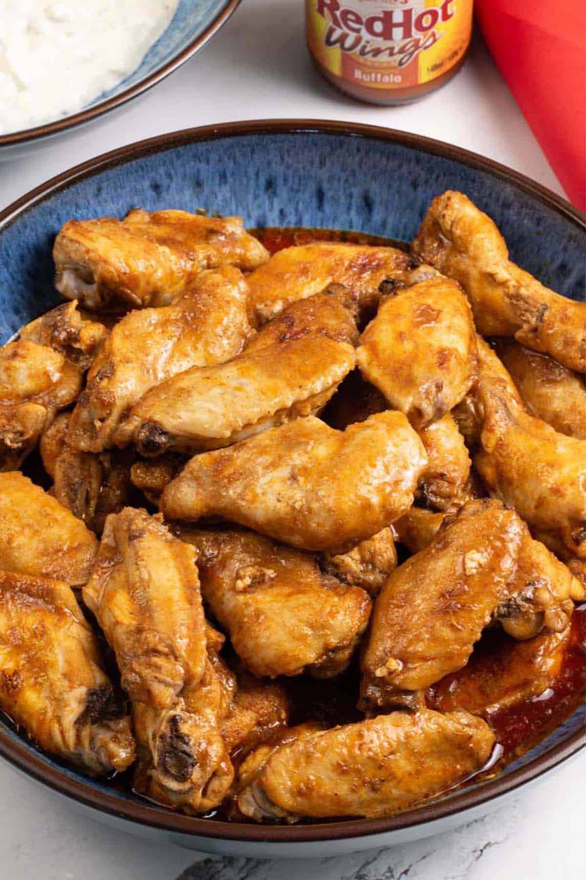 Buffalo chicken wings in a blue patterned bowl with a bowl of blue cheese dip, bottle of Frank's RedHot Wings Buffalo sauce and red napkin on the side.