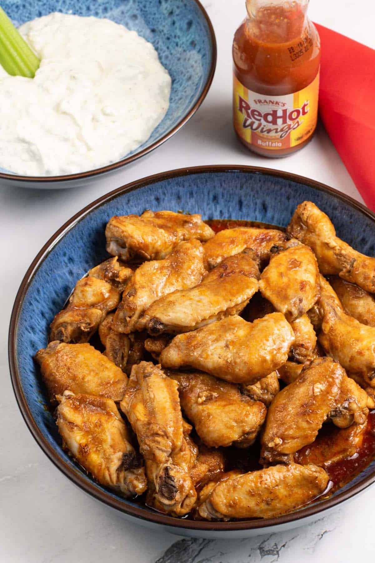 Buffalo chicken wings in a blue patterned bowl with a bowl of blue cheese dip, bottle of Frank's RedHot Wings Buffalo sauce and red napkin on the side.
