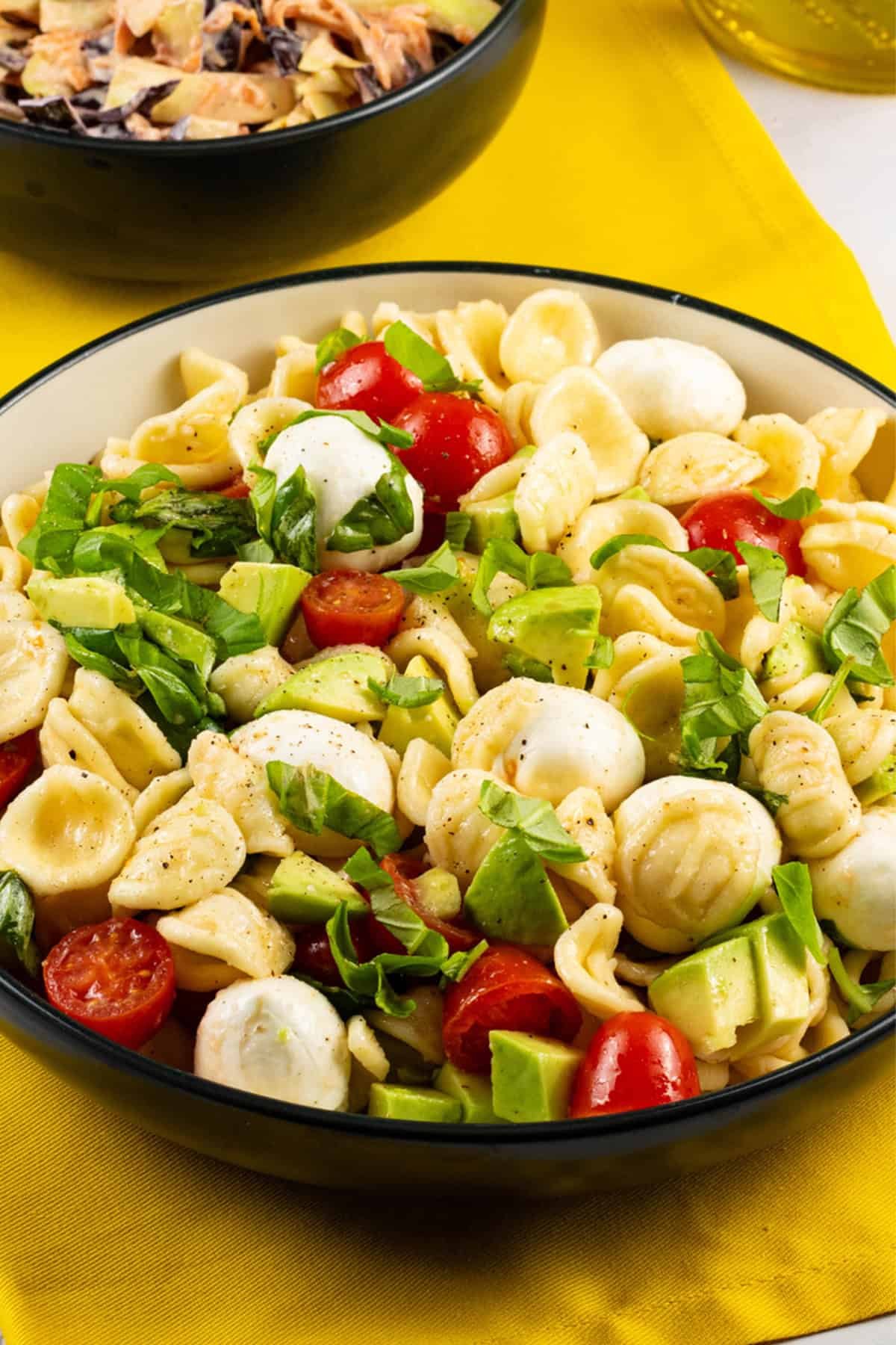 Caprese pasta salad in a black and cream bowl with a bowl of coleslaw and bottle of olive oil in the background.