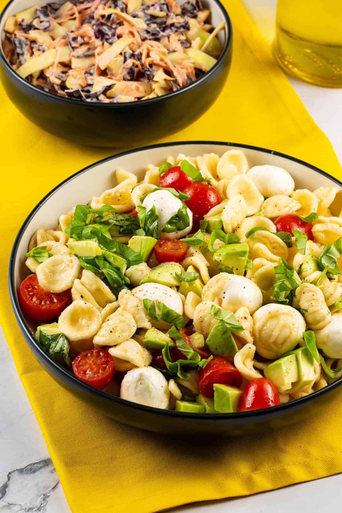 Caprese pasta salad in a black and cream bowl with a bowl of coleslaw and bottle of olive oil in the background.