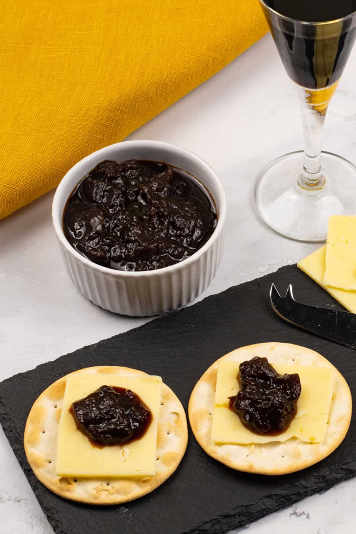 Caramelised red onion chutney made in the Thermomix in a white ramekin with crackers, cheese and chutney on a black slate, and a glass of port on the side.