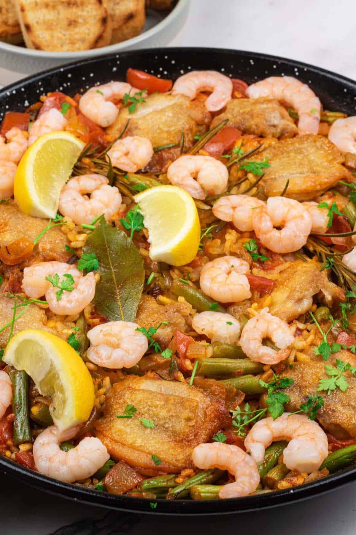 Chicken paella with prawns and beans in a paella pan with a dish of toasted crusty bread on the side.