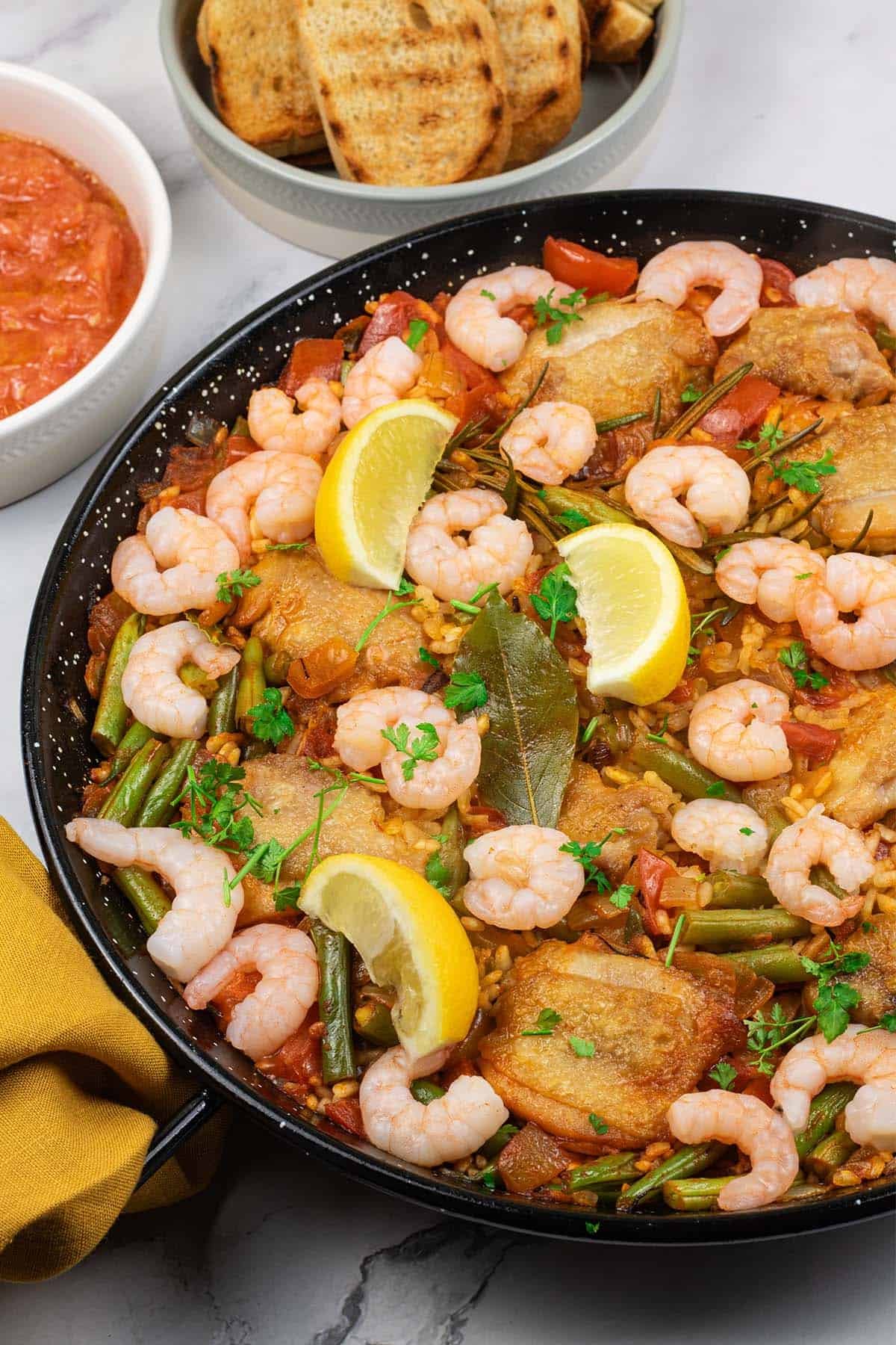 Chicken paella with prawns and beans in a paella pan with dishes of pan con tomate and toasted crusty bread on the side.