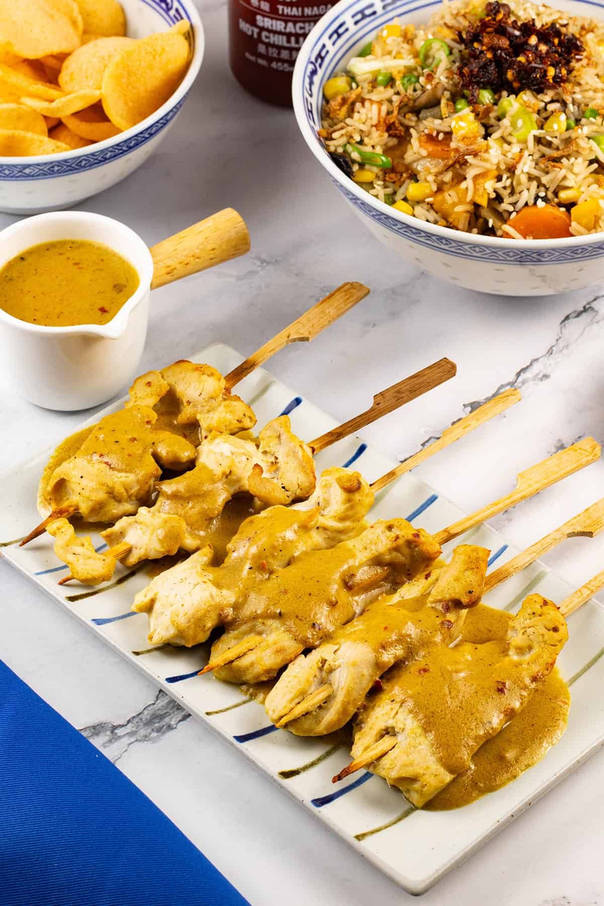 Chicken skewers with satay sauce on a striped platter, with a jug of sauce and bowls of vegetable fried rice and prawn crackers on the side.