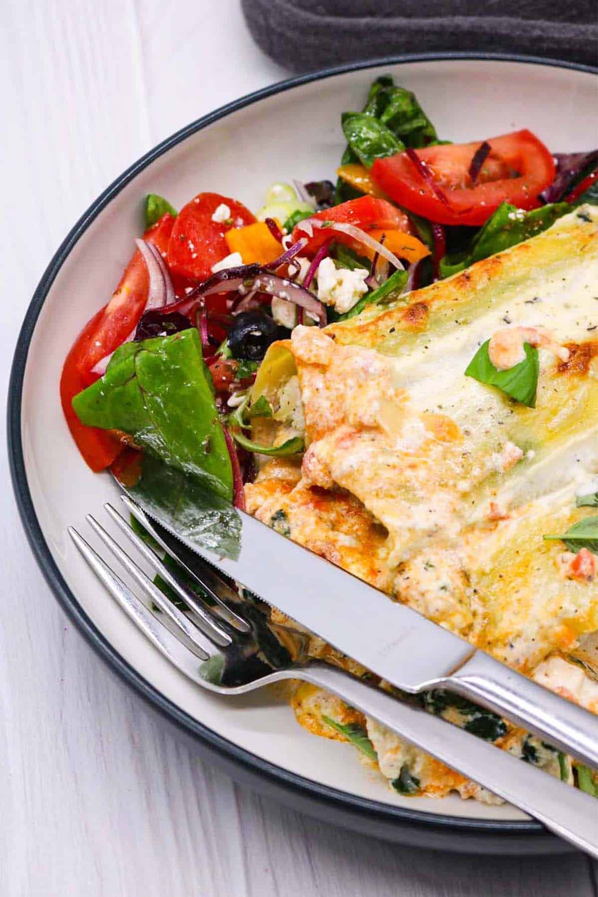 A portion of chicken, spinach and ricotta cannelloni in a bowl with salad, basil leaves and a fork and knife.