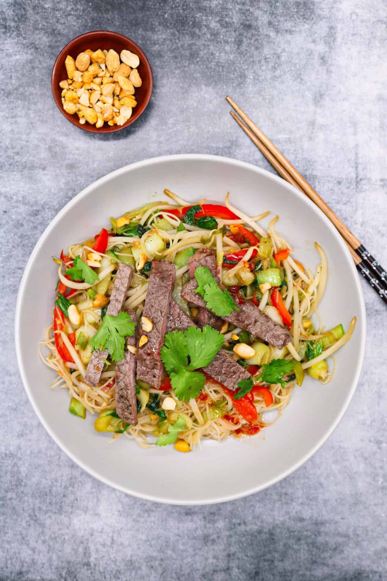 Chilli beef and vegetable rice noodles with peanuts in a grey bowl with chopsticks and a small dish of peanuts on the side.