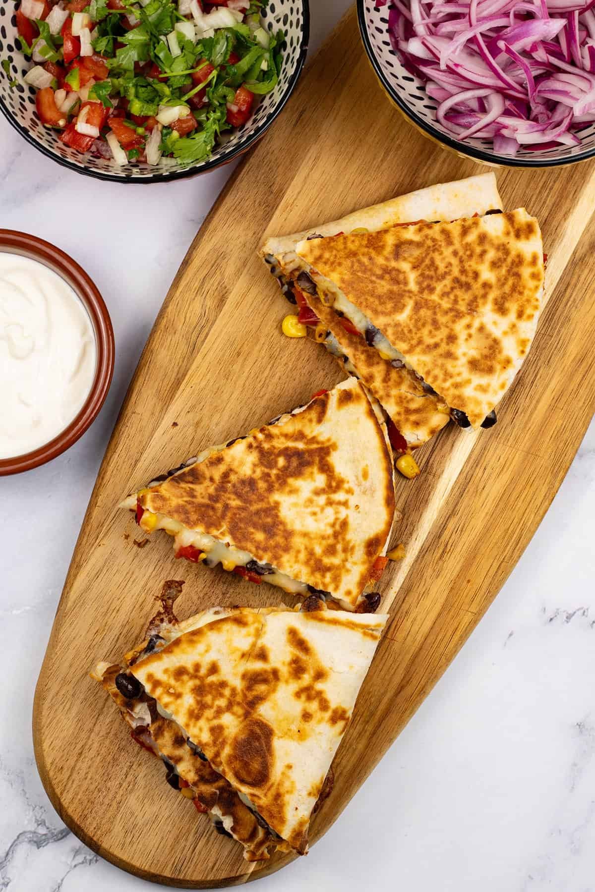2 portions of chipotle black bean and corn quesadillas on a board with bowls of pickled red onions, pico de gallo salsa and sour cream on the side.