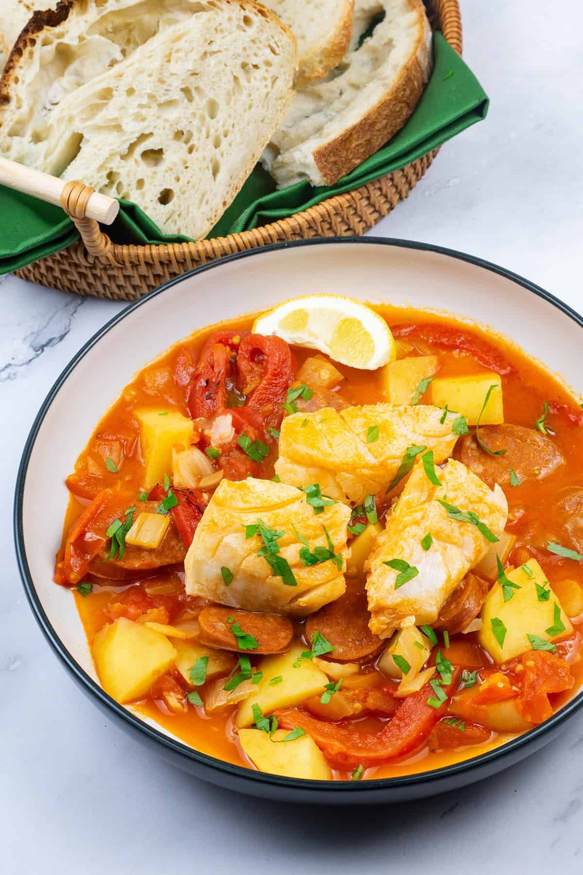 A portion of cod and chorizo stew in a bowl with a basket of crusty bread on the side.