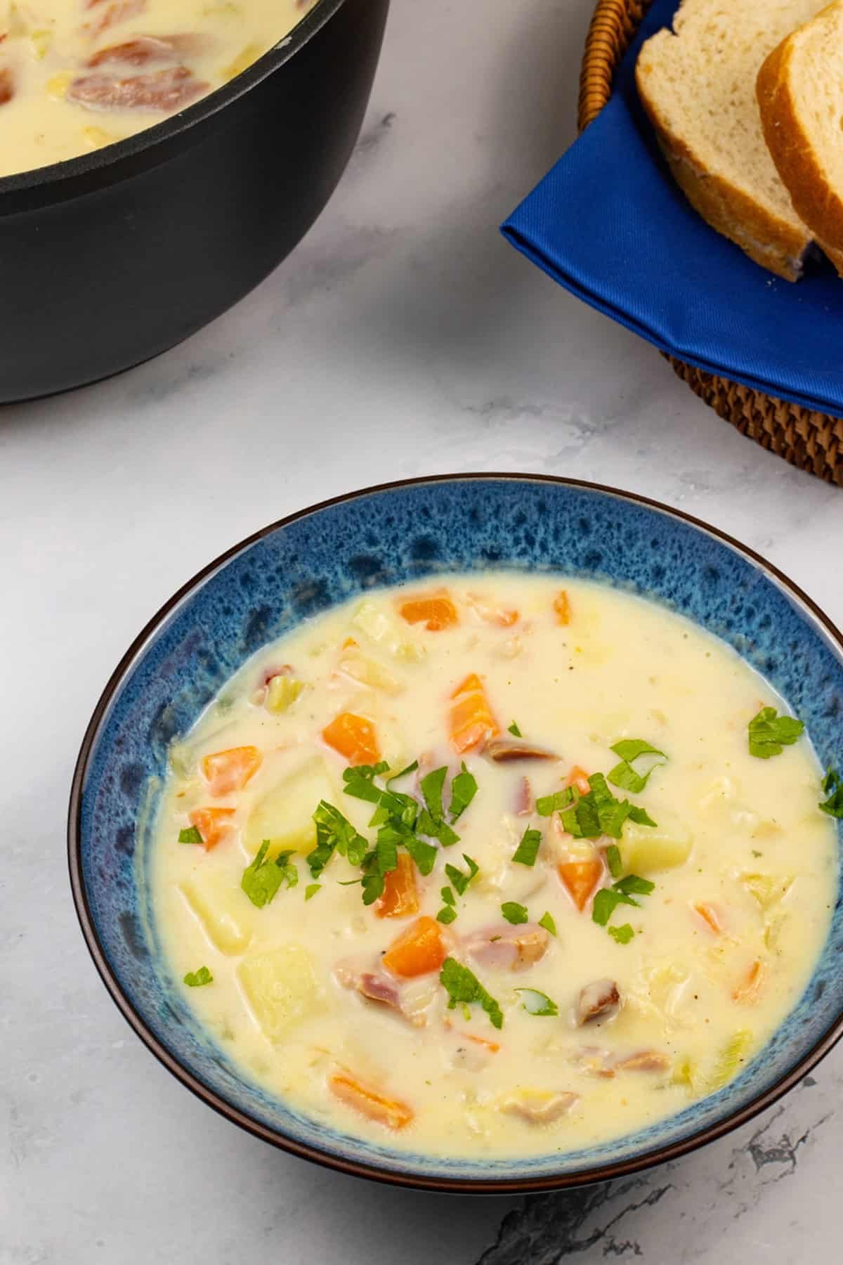 Creamy ham and potato soup in a blue patterened bowl with crusty bread and pot of soup on the side.