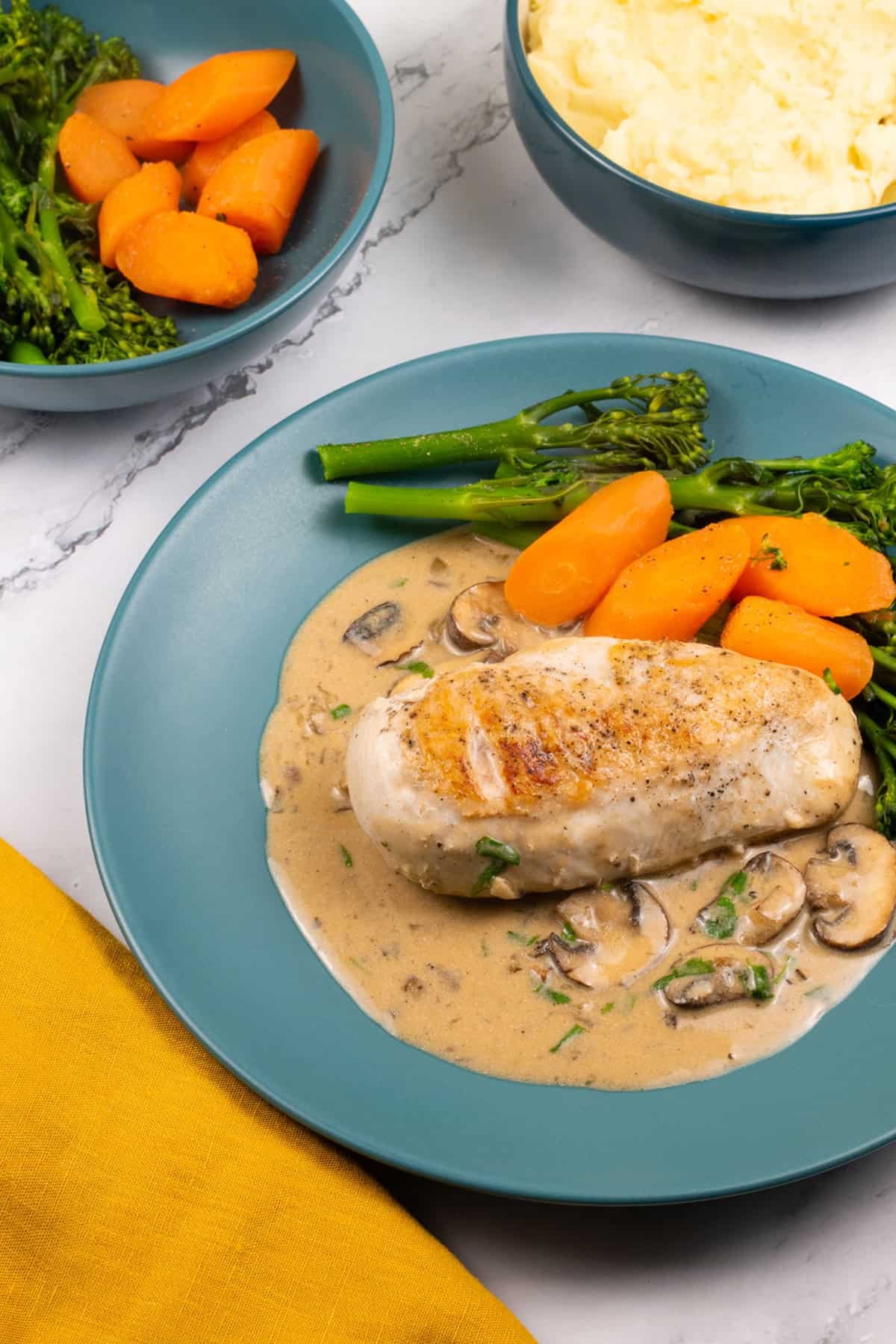 A portion of creamy mushroom chicken with broccoli and carrots on a teal dinner plate with bowls of broccoli and carrots, and mashed potatoes on the side.