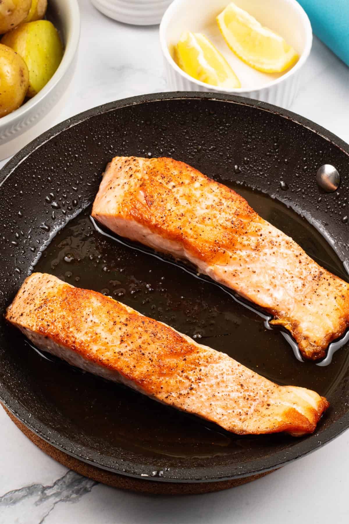 Crispy pan-fried salmon fillets in the frying pan with lemon wedges and baby potatoes on the side.