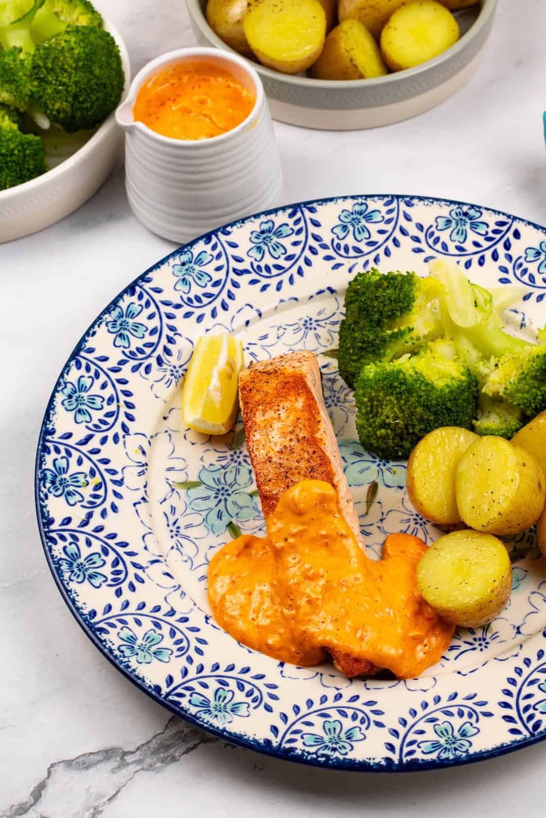 A crispy pan-fried salmon fillet plated with red pepper and paprika hollandaise, baby potatoes, broccoli and a lemon wedge.