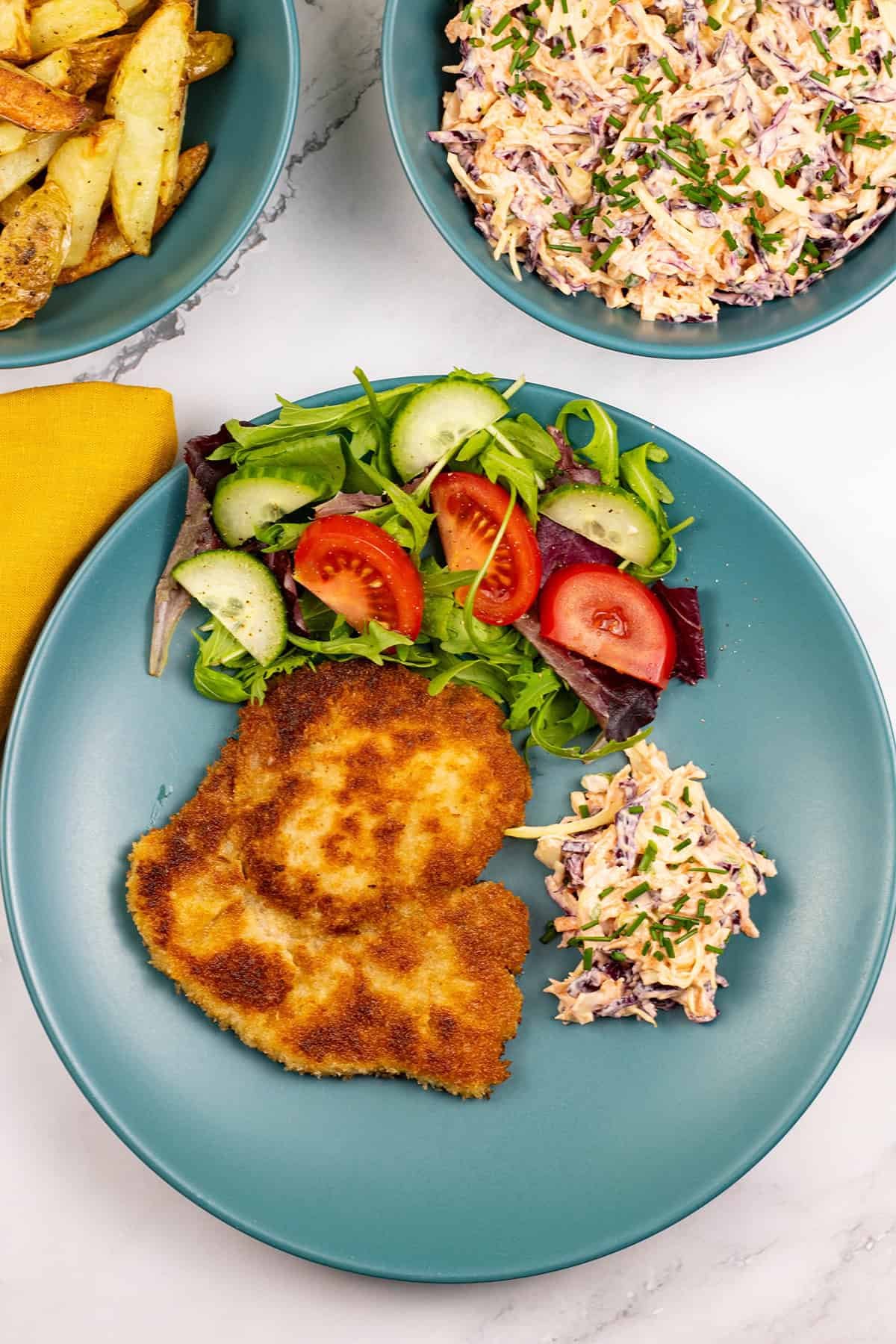 A crispy pork schnitzel, coleslaw and salad on a teal dinner plate, with bowls of chips and coleslaw on the side.