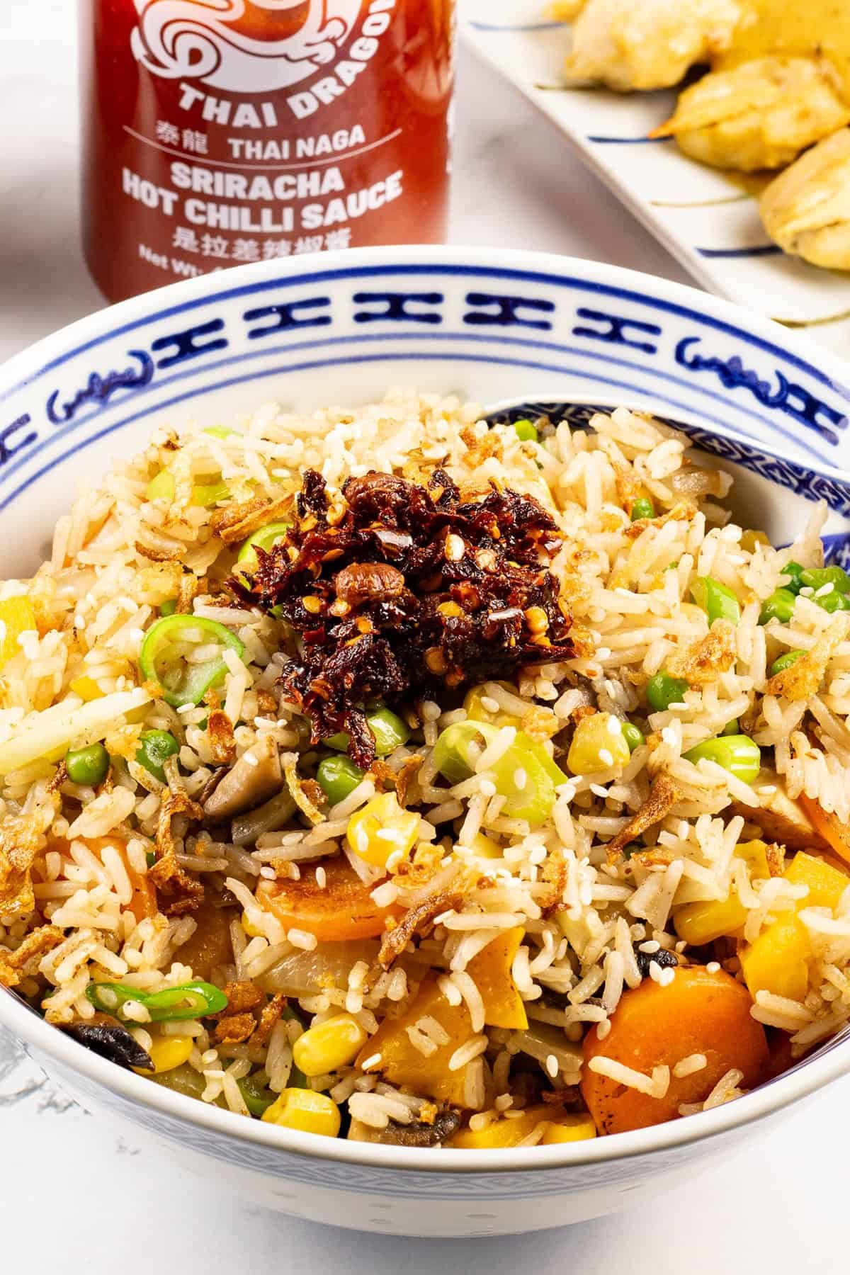 Close up of easy vegetable fried rice in a blue and white oriental serving bowl with spoon and a bottle of sriracha on the side.
