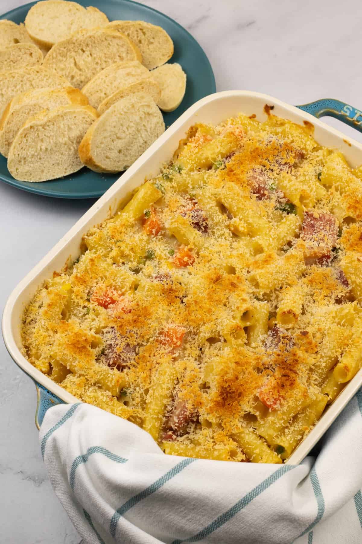 Ham and vegetable pasta bake in a rectangle Staub dish with a plate of crusty bread and a teal and white striped towel on the side.