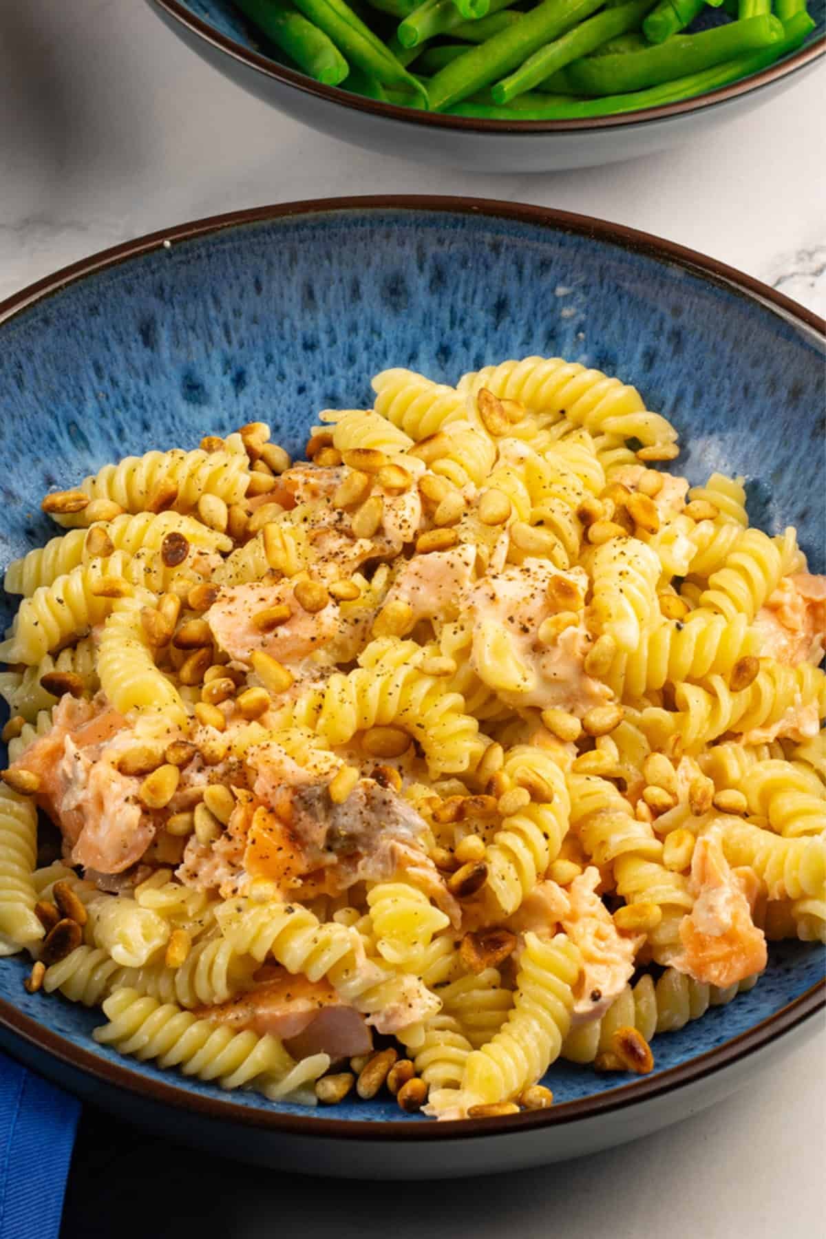 Hot smoked salmon with creamy pasta and pine nuts in a blue patterned bowl, with a bowl of green beans on the side.