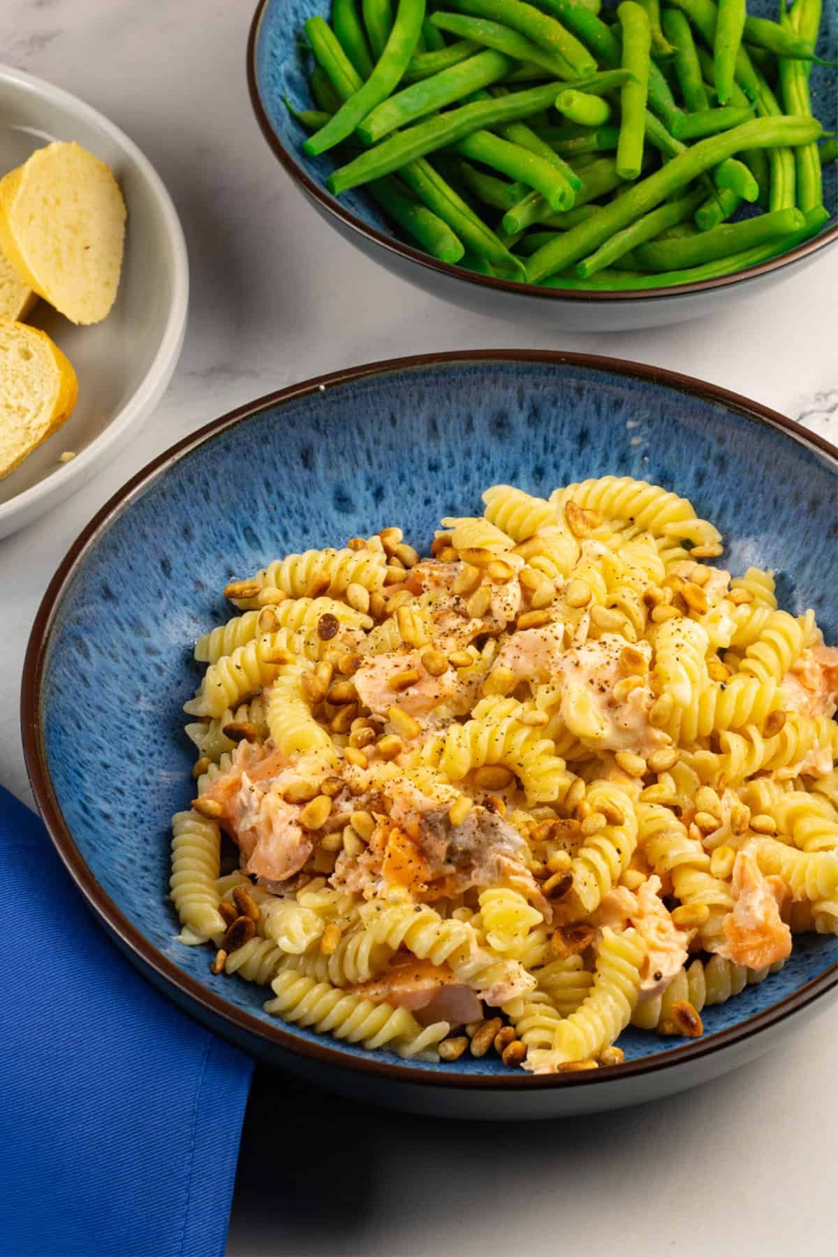 Hot smoked salmon with creamy pasta and pine nuts in a blue patterned bowl, with a bowl of green beans and plate of crusty bread on the side.