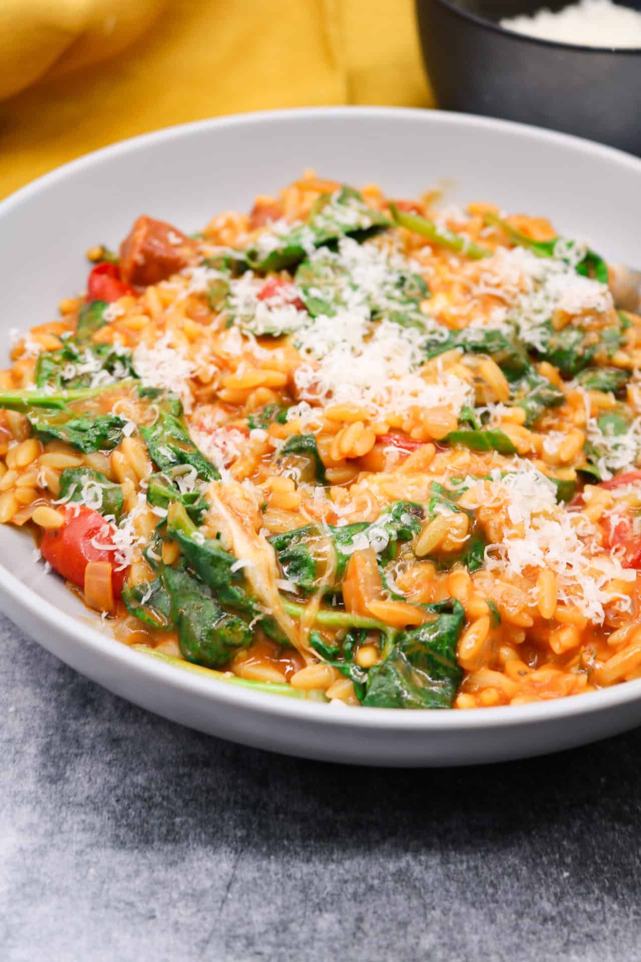 Orzo risotto with chorizo and baby spinach in a grey bowl with fork and small bowl of parmesan and mustard napkin on the side.