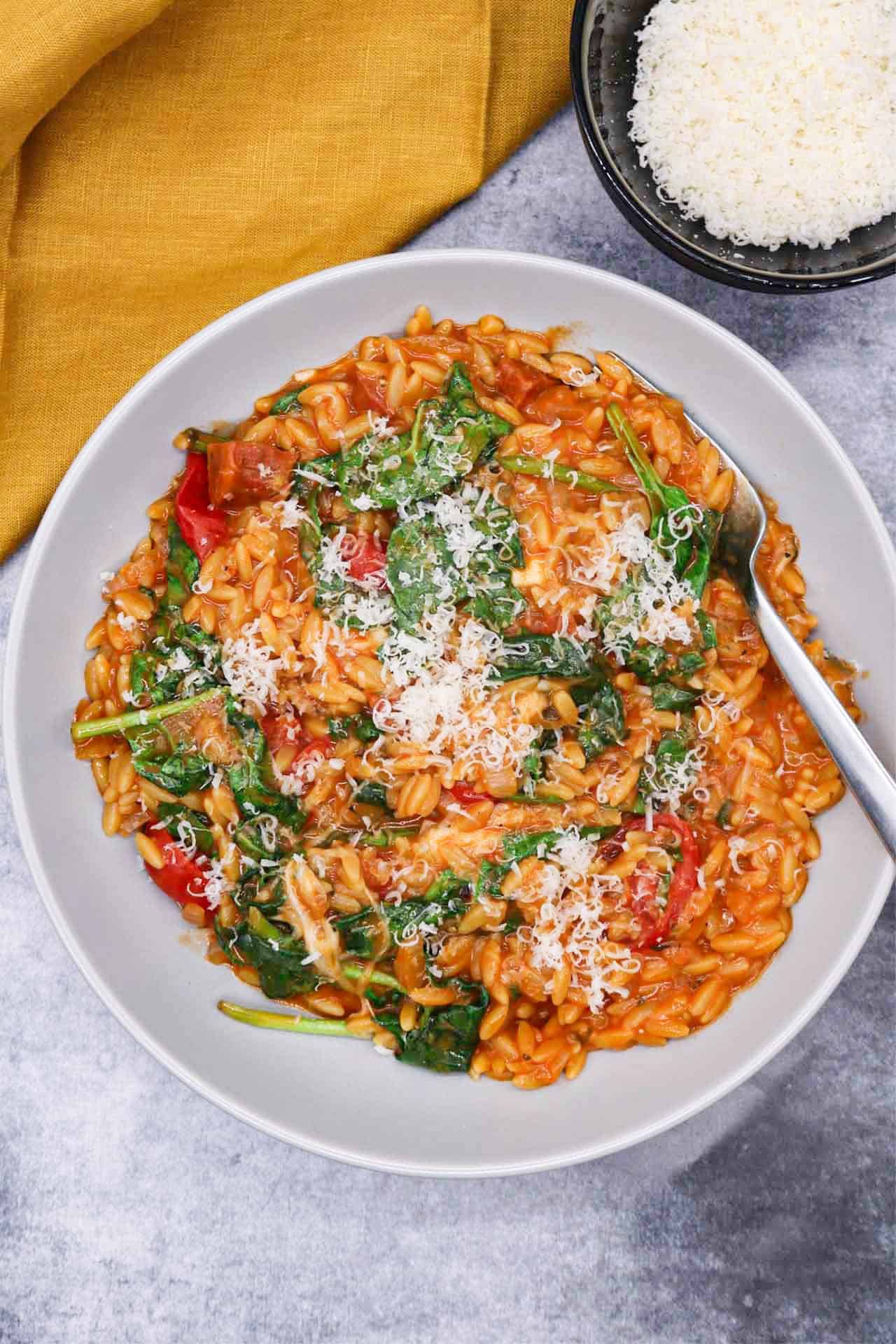 Orzo risotto with chorizo and baby spinach in a grey bowl with fork and small bowl of parmesan and mustard napkin on the side.