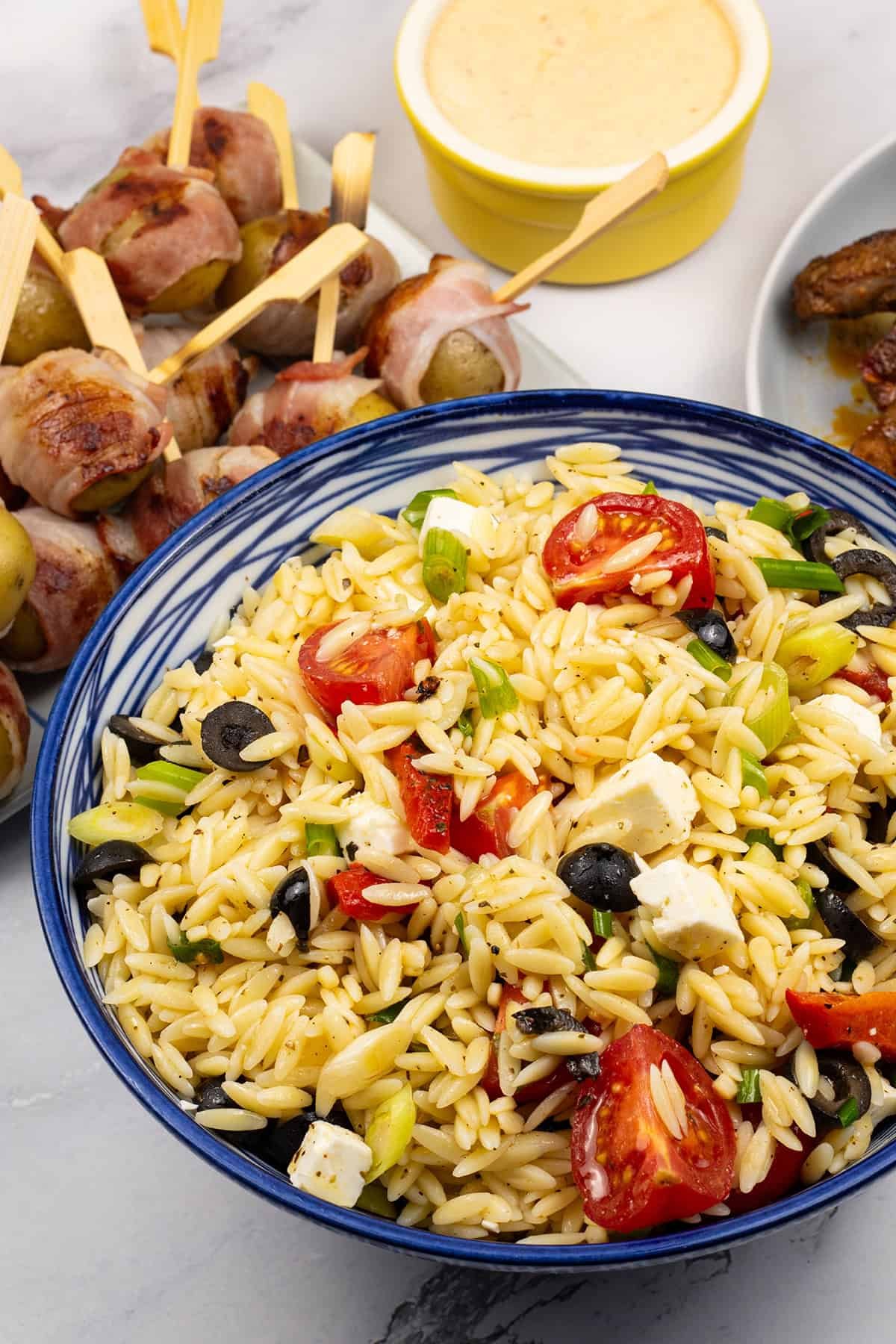 Orzo salad with feta in a blue and white striped bowl with a platter of baby new potatoes wrapped in bacon and a ramekin of harissa cheese dip on the side.