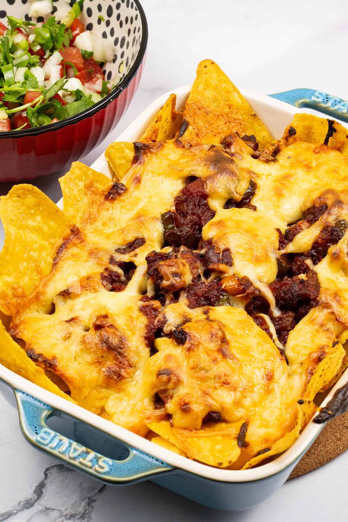 Party nachos with chorizo and spicy cheese sauce in a small rectangular dish with a bowl of pico de gallo salsa on the side.