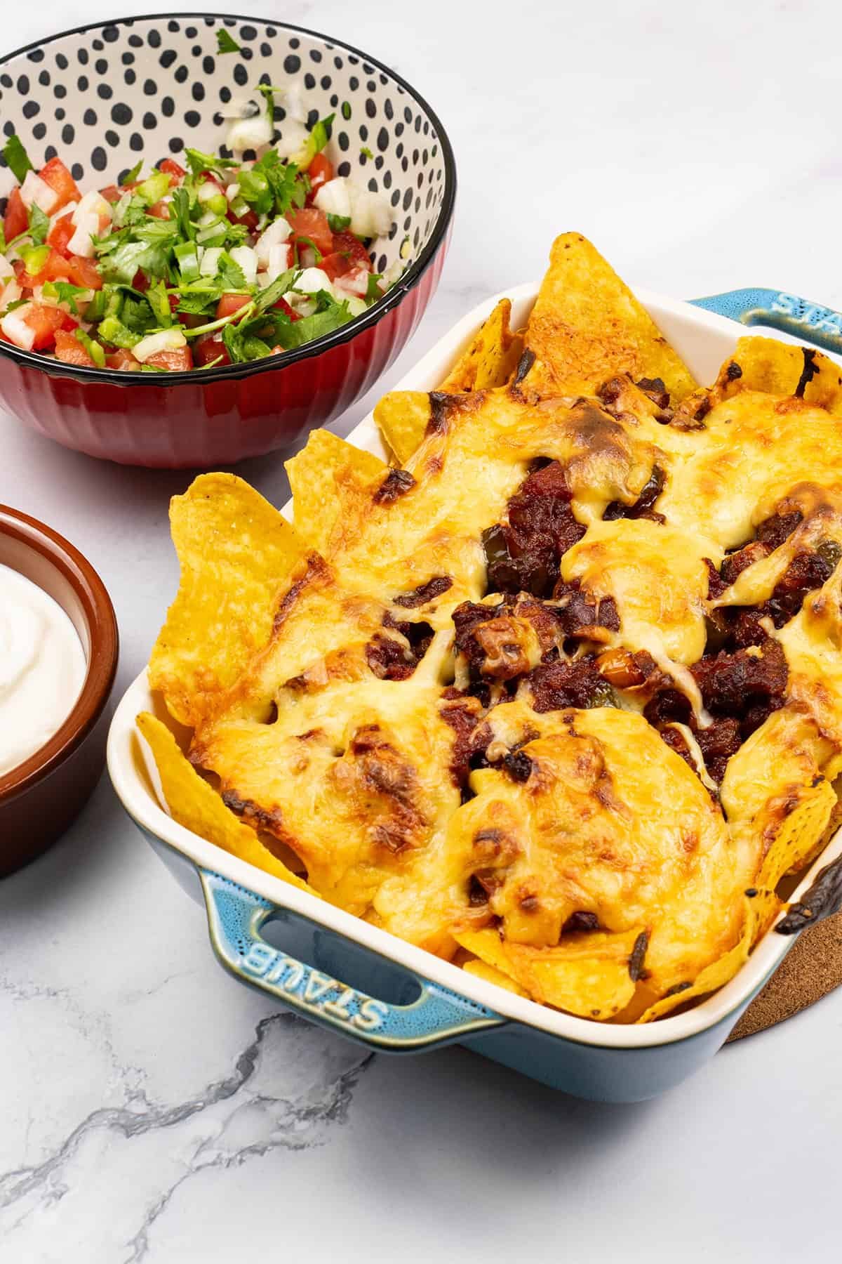Party nachos with chorizo and spicy cheese sauce in a small rectangular dish with bowls of pico de gallo salsa and sour cream on the side.