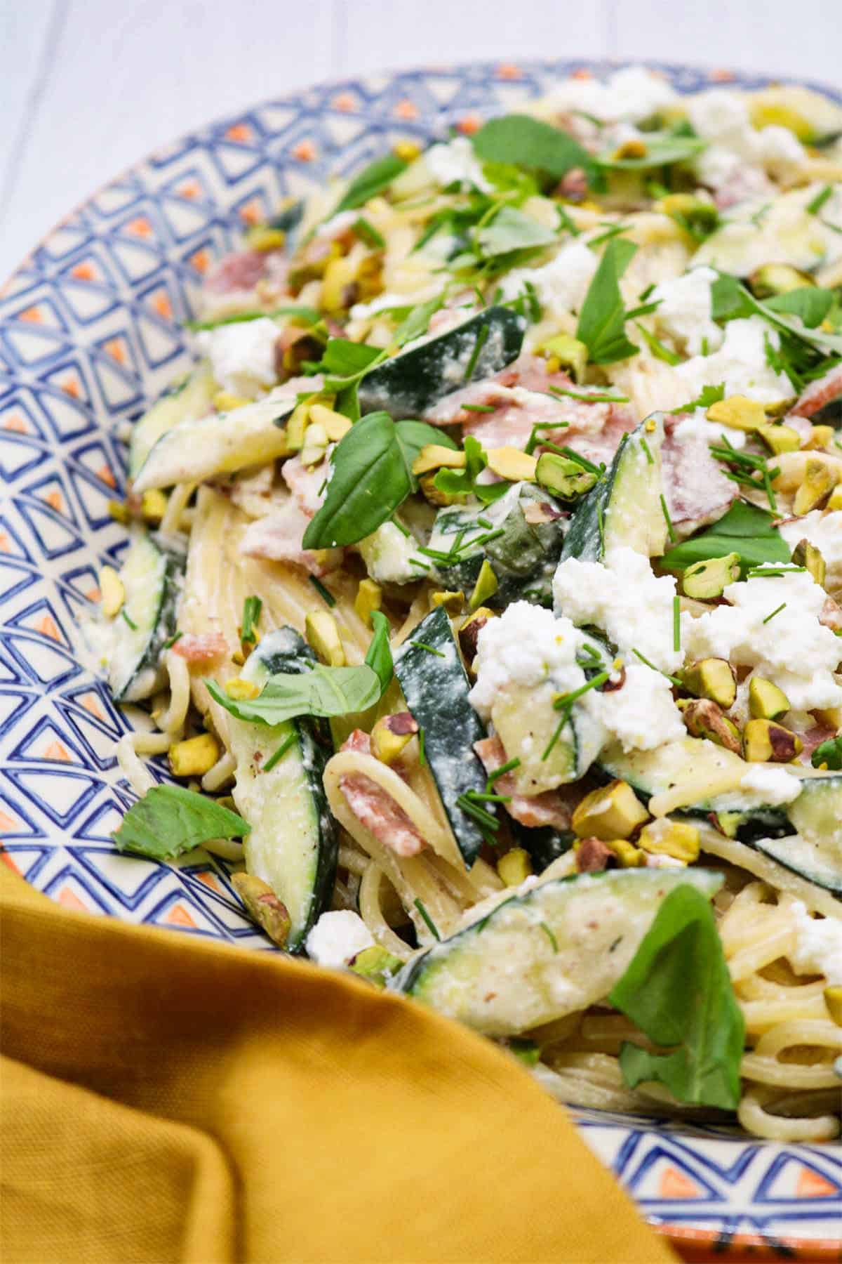 Pasta with crispy bacon, courgette and herbs on an orange, blue and white patterned oval platter.