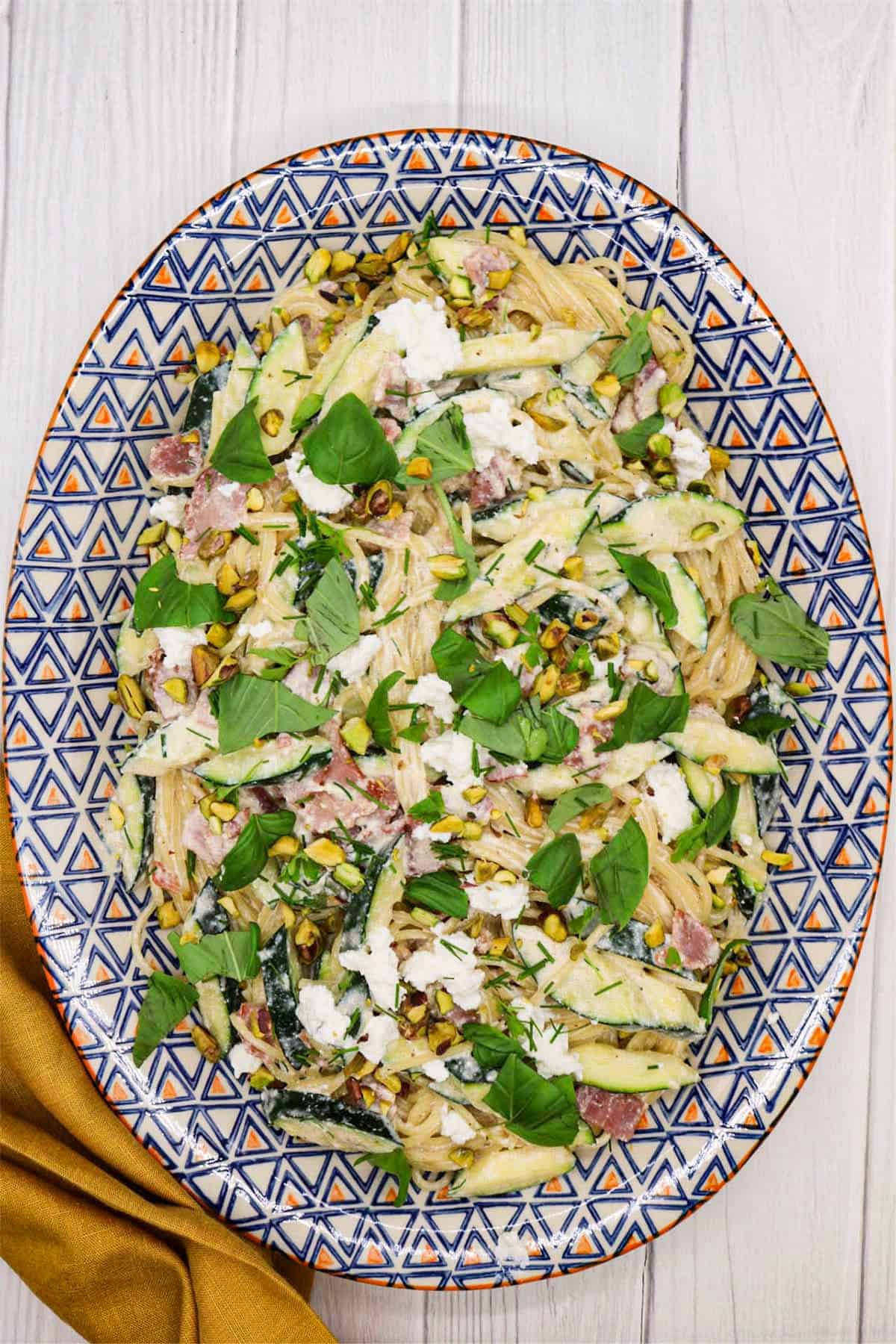 Pasta with crispy bacon, courgette and herbs on an orange, blue and white patterned oval platter.