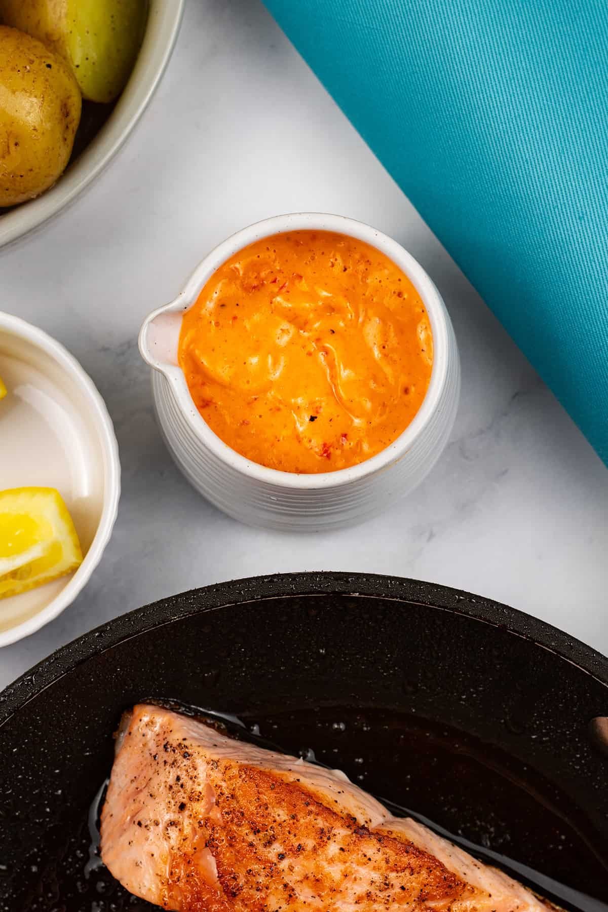 Red pepper and paprika hollandaise sauce made in the Thermomix in a small white jug. Crispy salmon fillet, lemon wedges and baby potatoes on the side.