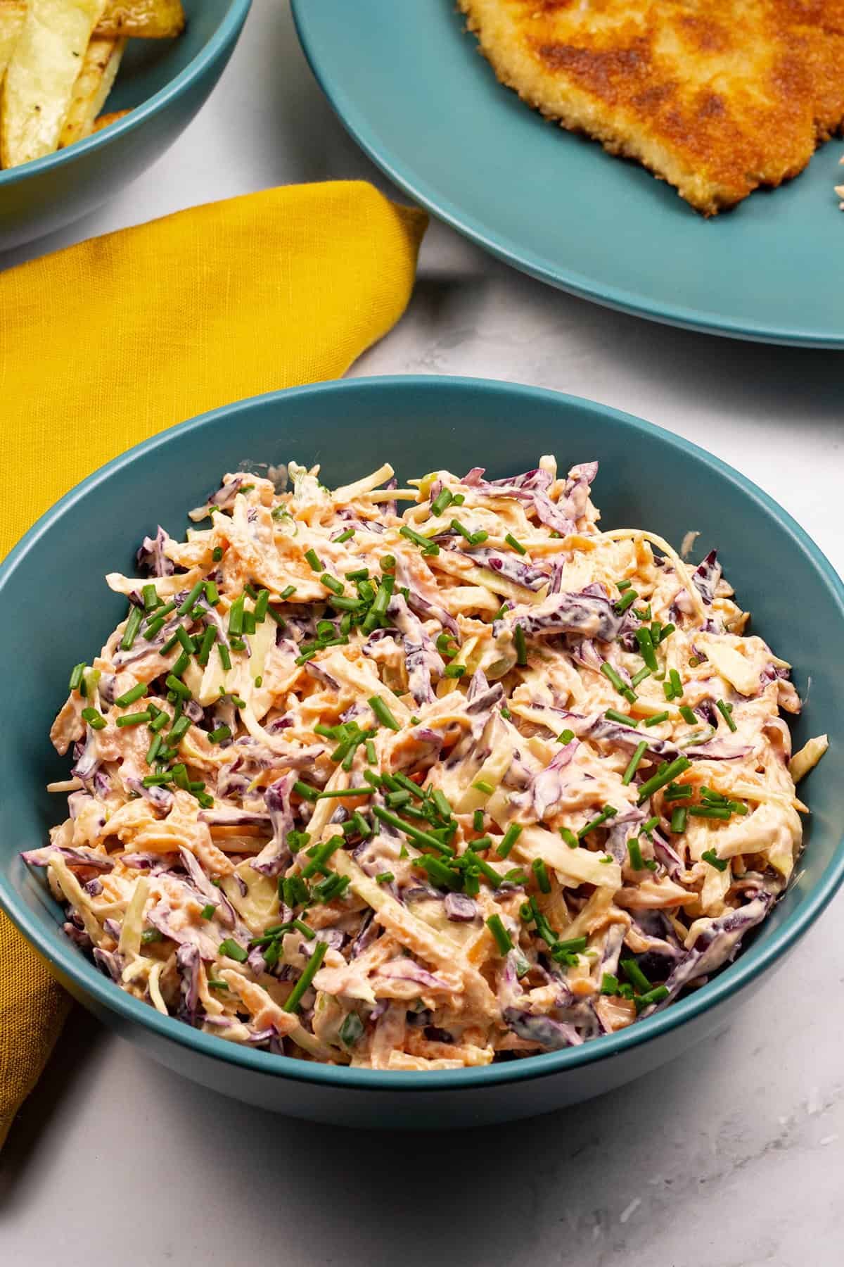 Red and white coleslaw in a teal bowl with a mustard napkin, bowl of chips and dinner plate with pork schnitzel on the side.