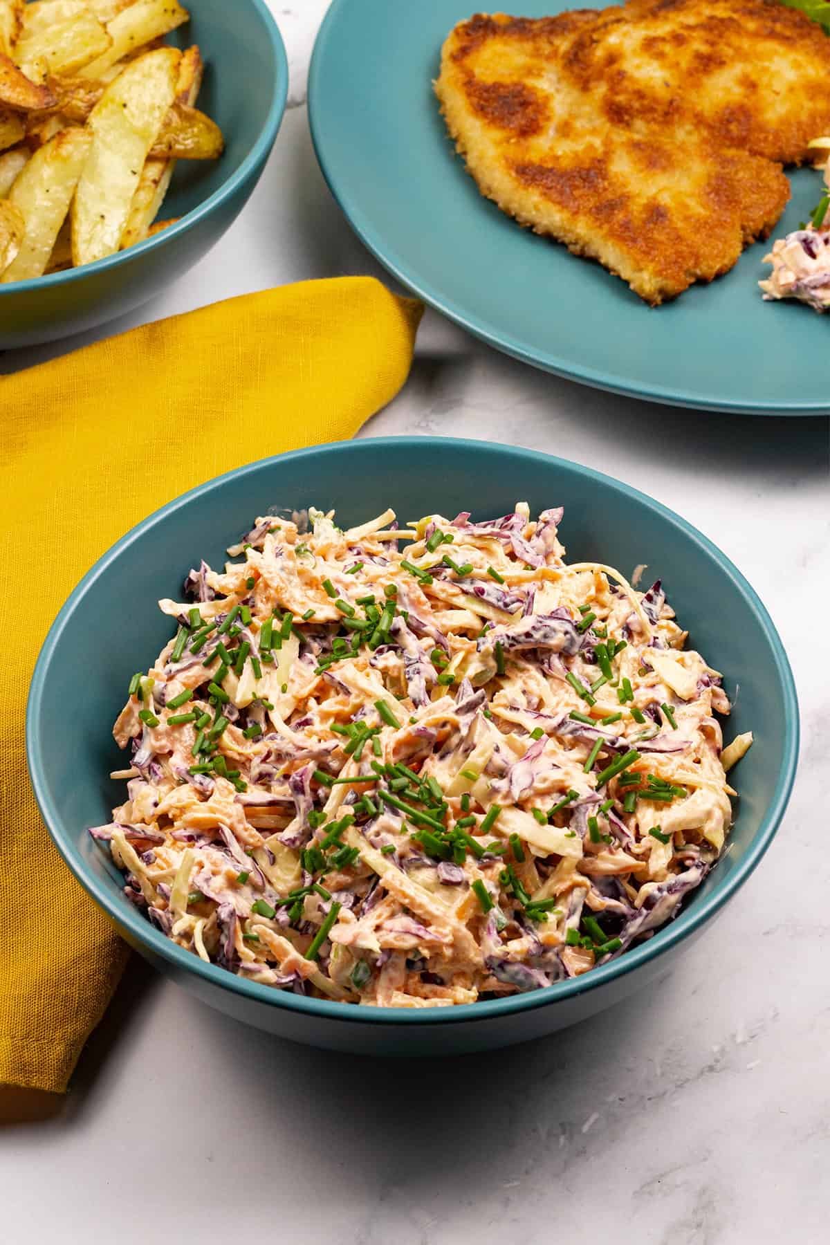 Red and white coleslaw in a teal bowl with a mustard napkin, bowl of chips and dinner plate with pork schnitzel on the side.