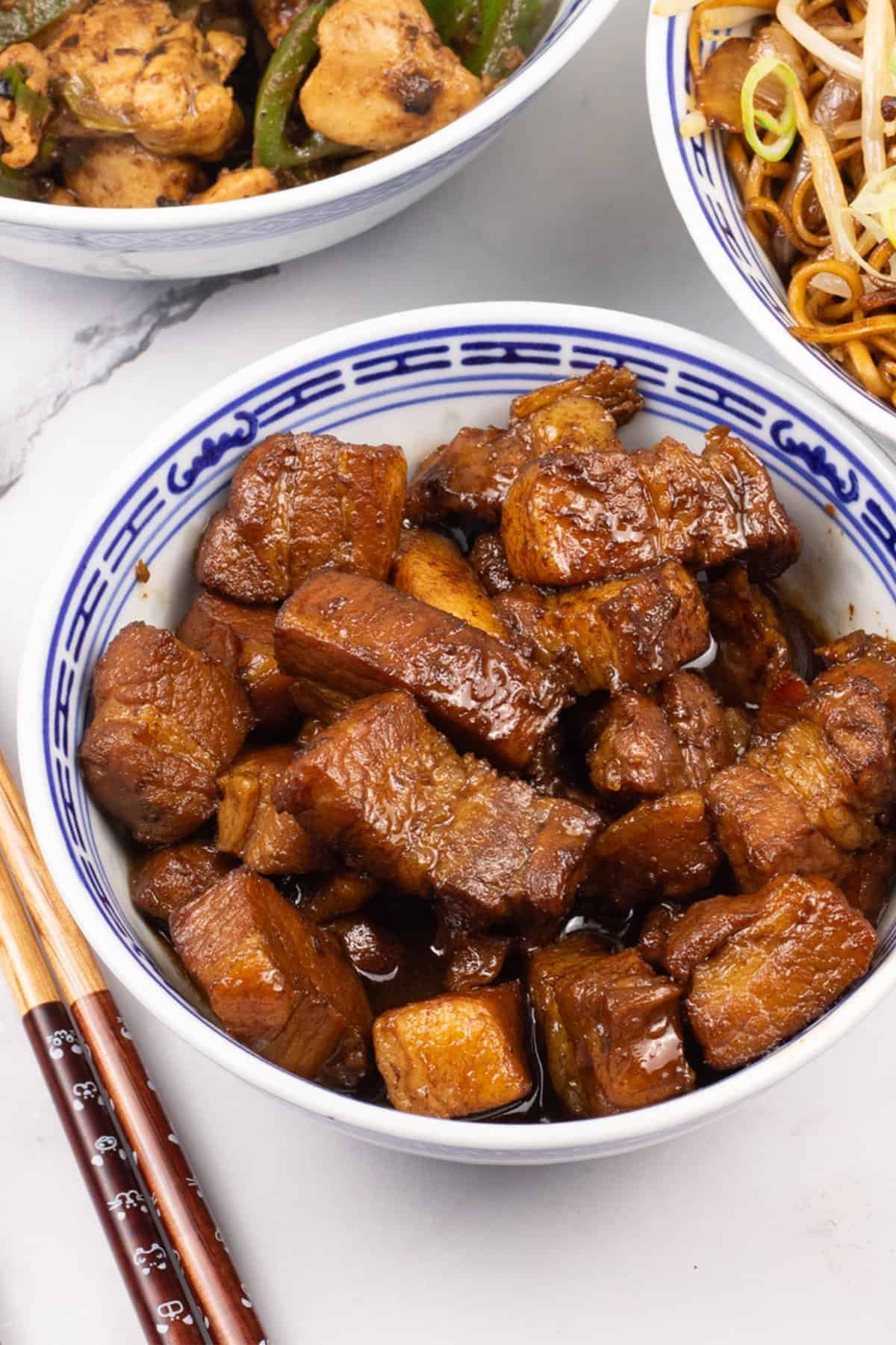 Shanghai-style braised pork belly in an oriental bowl with bowls of chicken and green peppers and fried noodles, and chopsticks on the side.