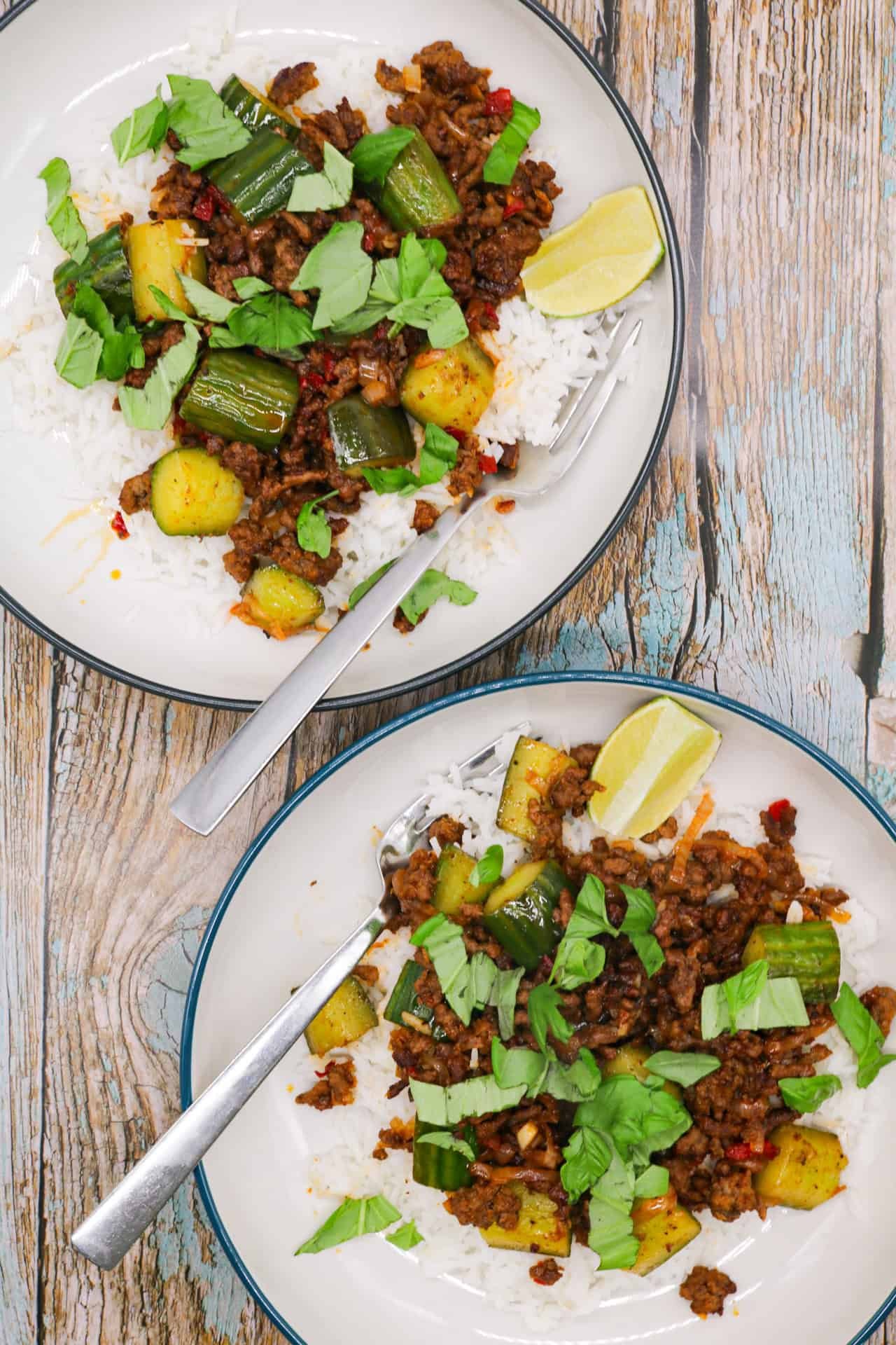 Two bowls of spicy beef and crushed cucumber stir-fry served on rice with forks and lime wedges.