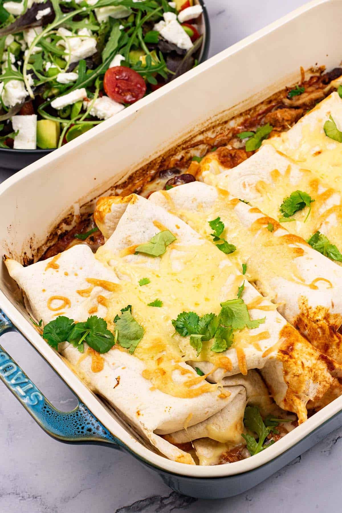 Turkey enchiladas in a large rectangle oven dish with a bowl of salad on the side.