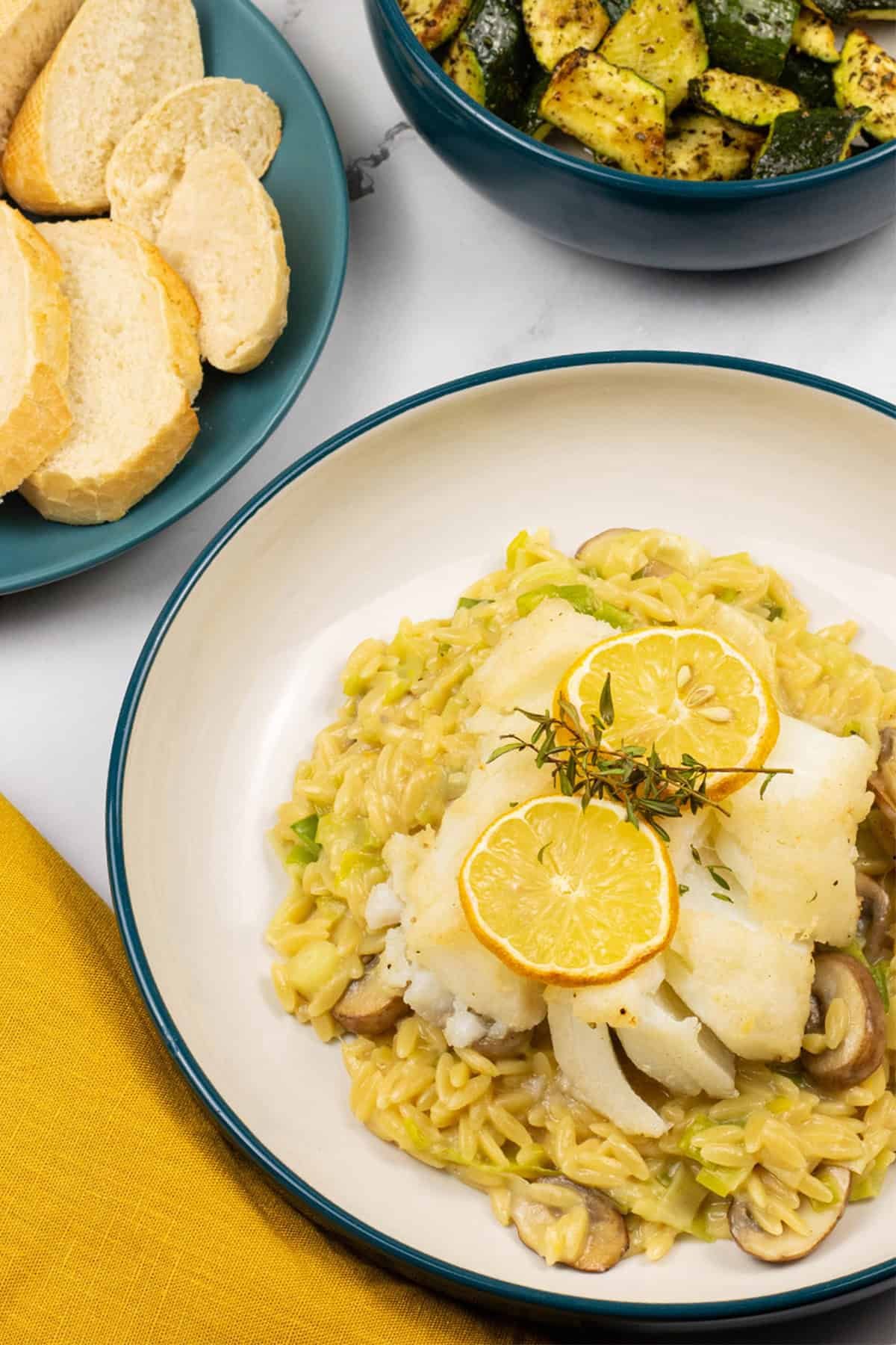 Baked cod with creamy mushroom and leek orzo in a bowl with a plate of sliced baguette and a bowl of air fryer courgettes on the side.