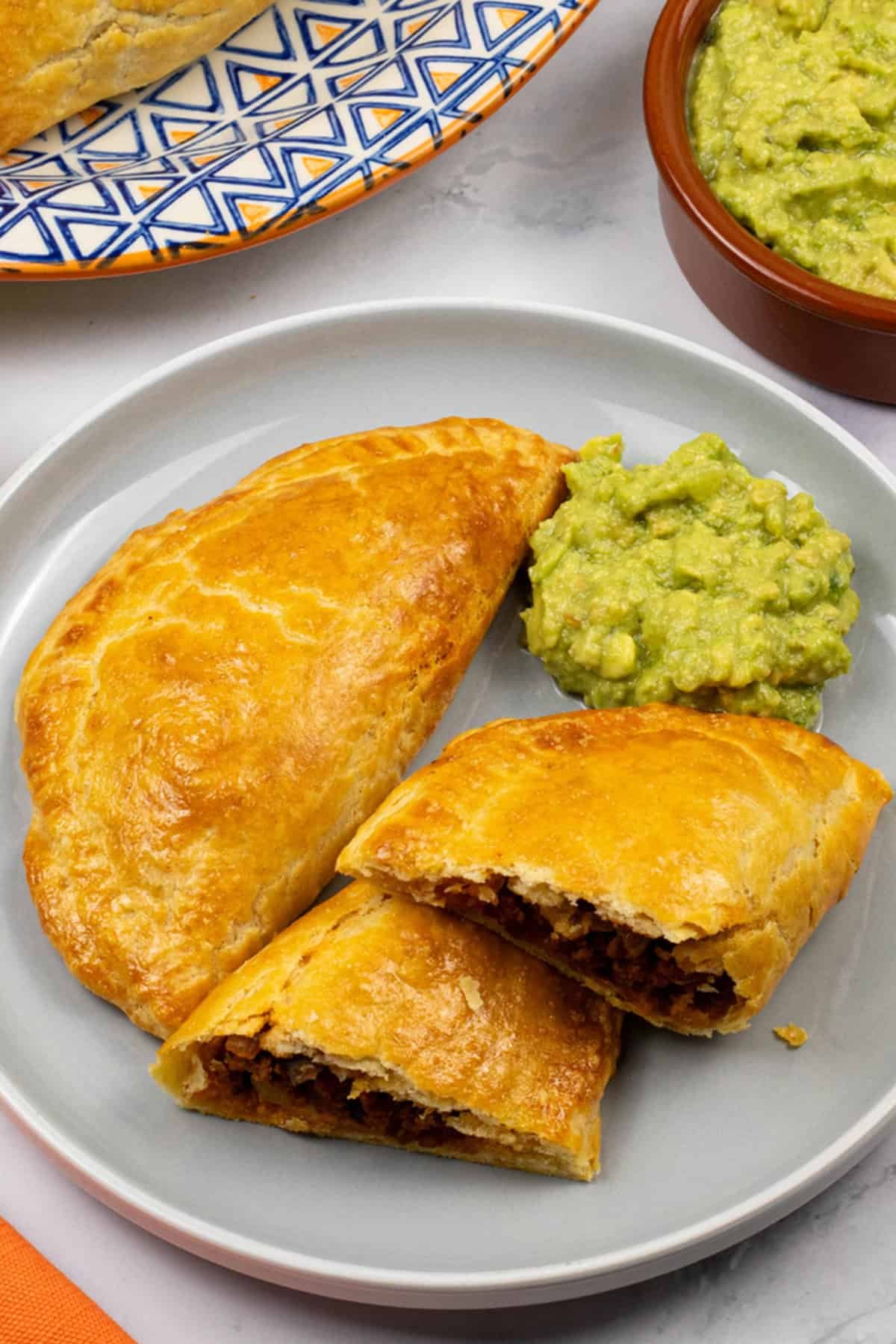 2 beef and chorizo empanadas on a grey side plate with some guacamole, a platter of empanadas and dish of guacamole on the side.