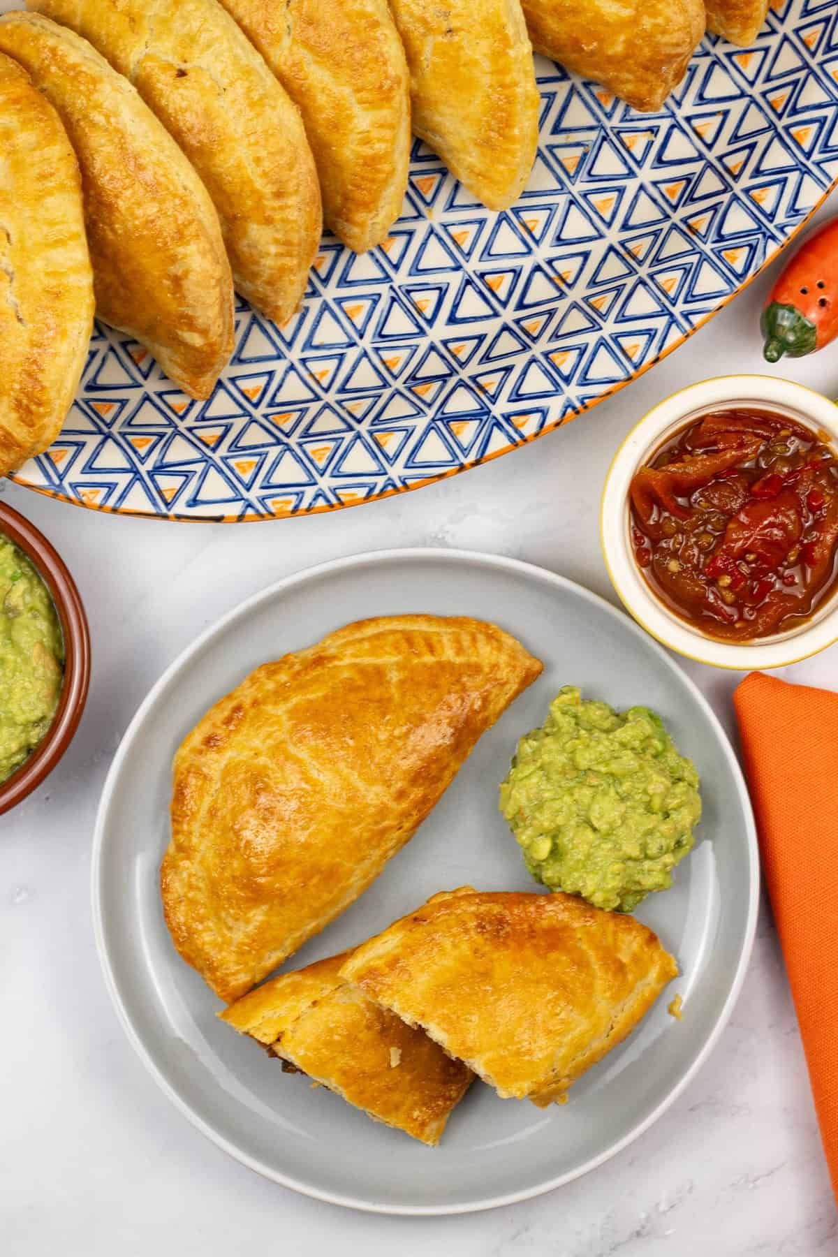 2 beef and chorizo empanadas on a grey side plate with some guacamole, a platter of empanadas and dishes of guacamole and chilli and tomato jam on the side.