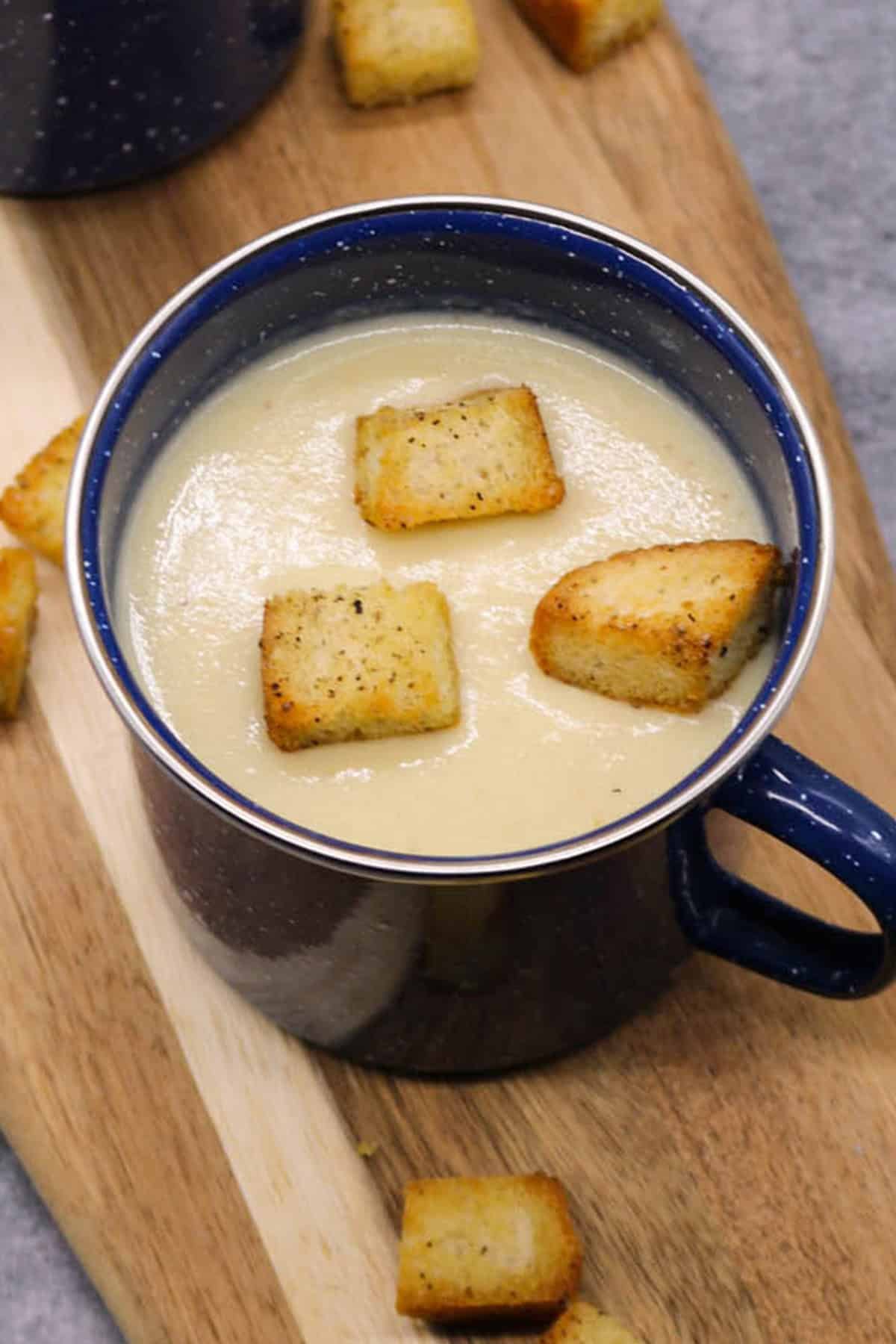 Cauliflower cheese soup in a blue mug sitting on a board with croutons on the side.