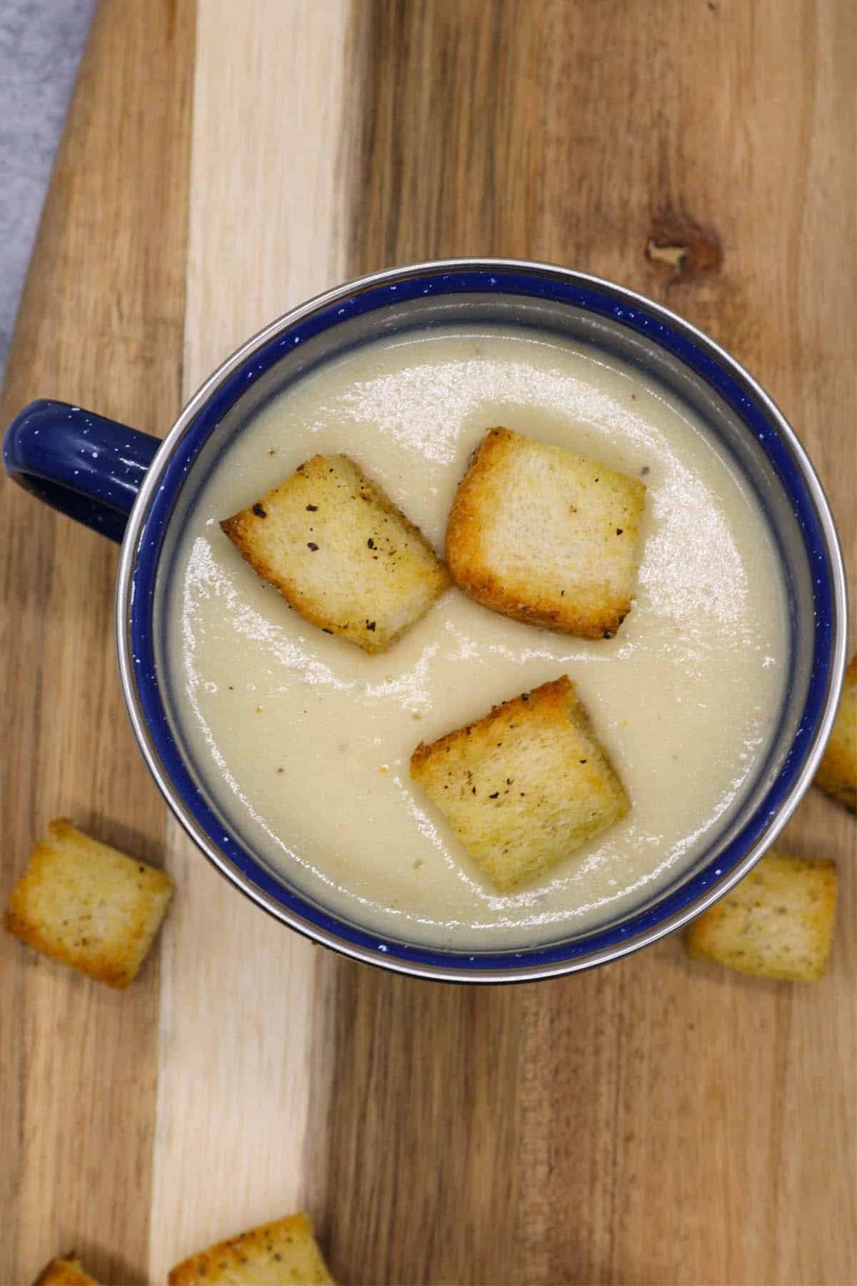 Cauliflower cheese soup in a blue mug sitting on a board with croutons on the side.
